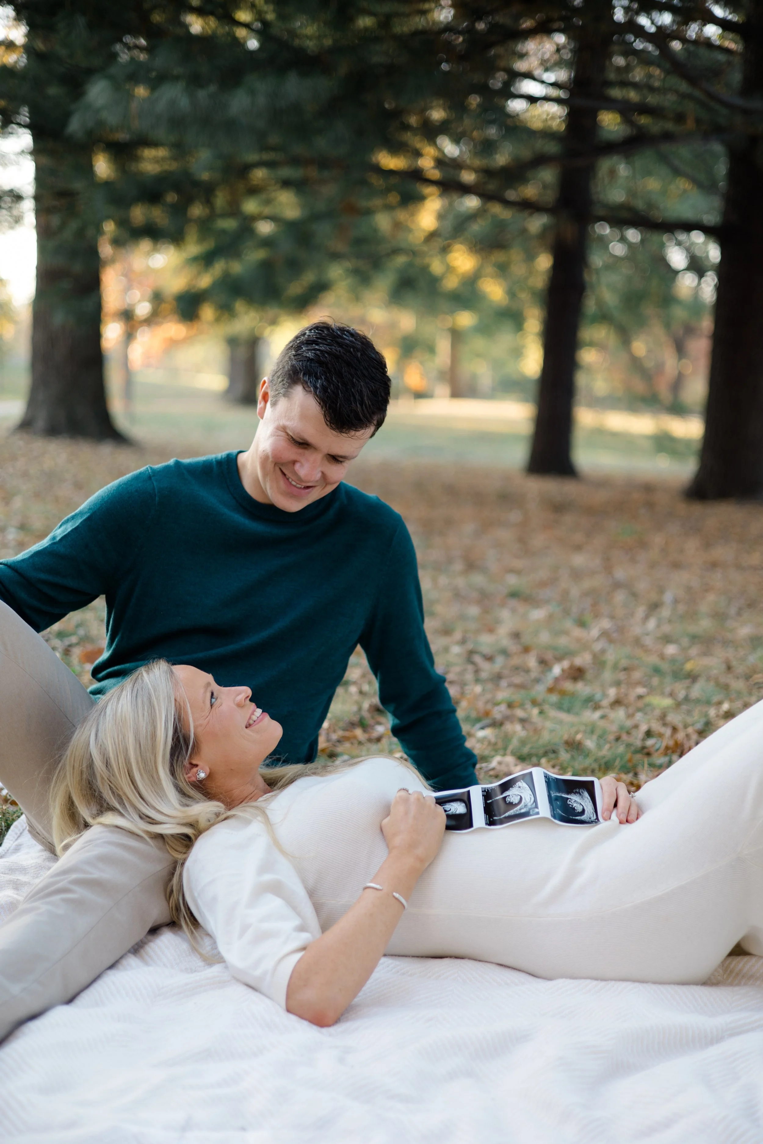 A woman with blonde hair lying on her back in bed with a pregnancy ultrasound printout on her stomach, smiling at her partner, a man with dark hair, who is standing and leaning over her, in a park with trees in the background.