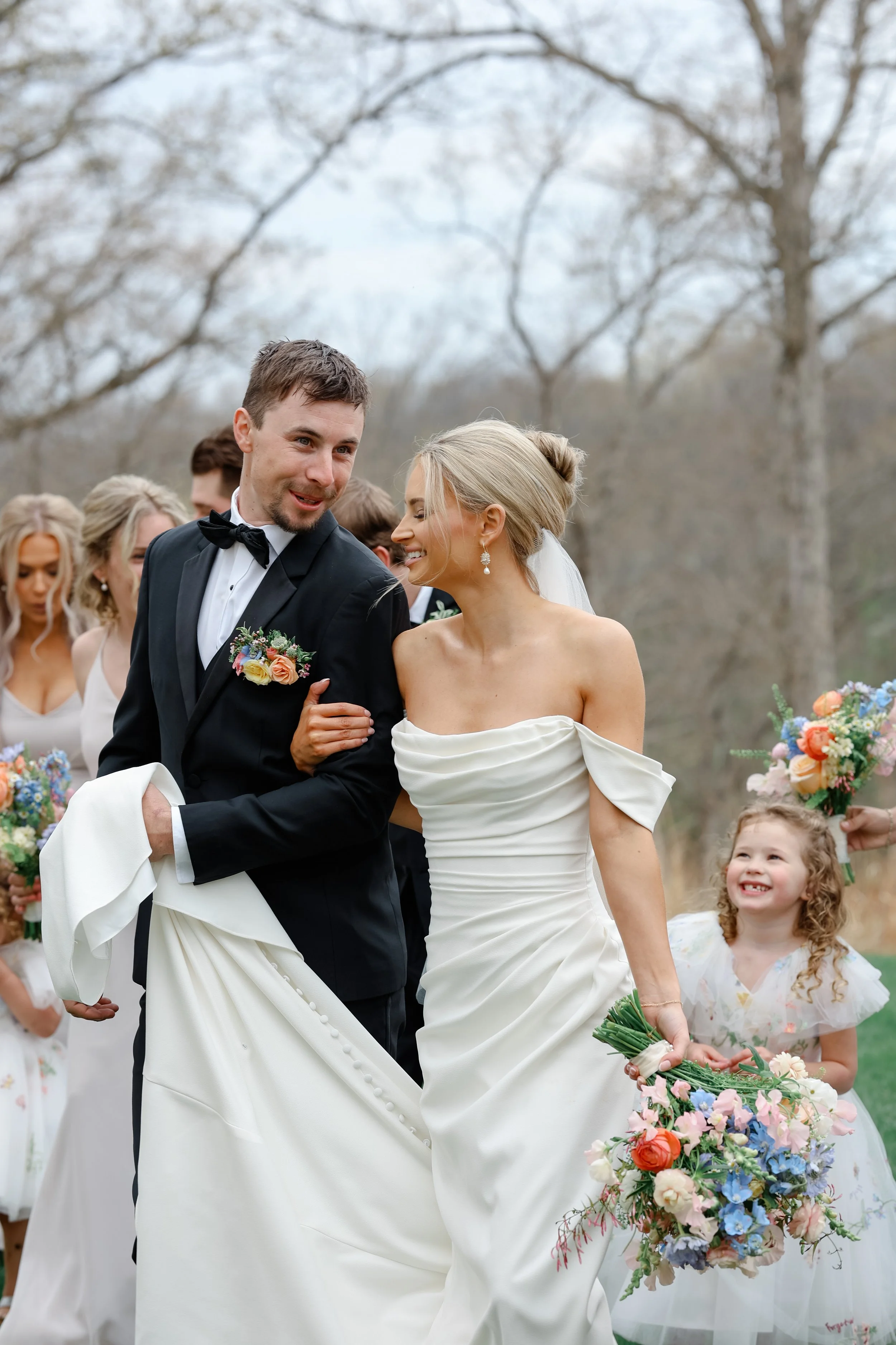 A bride and groom smiling and looking at each other during their outdoor wedding ceremony, surrounded by guests and children, with a background of leafless trees.