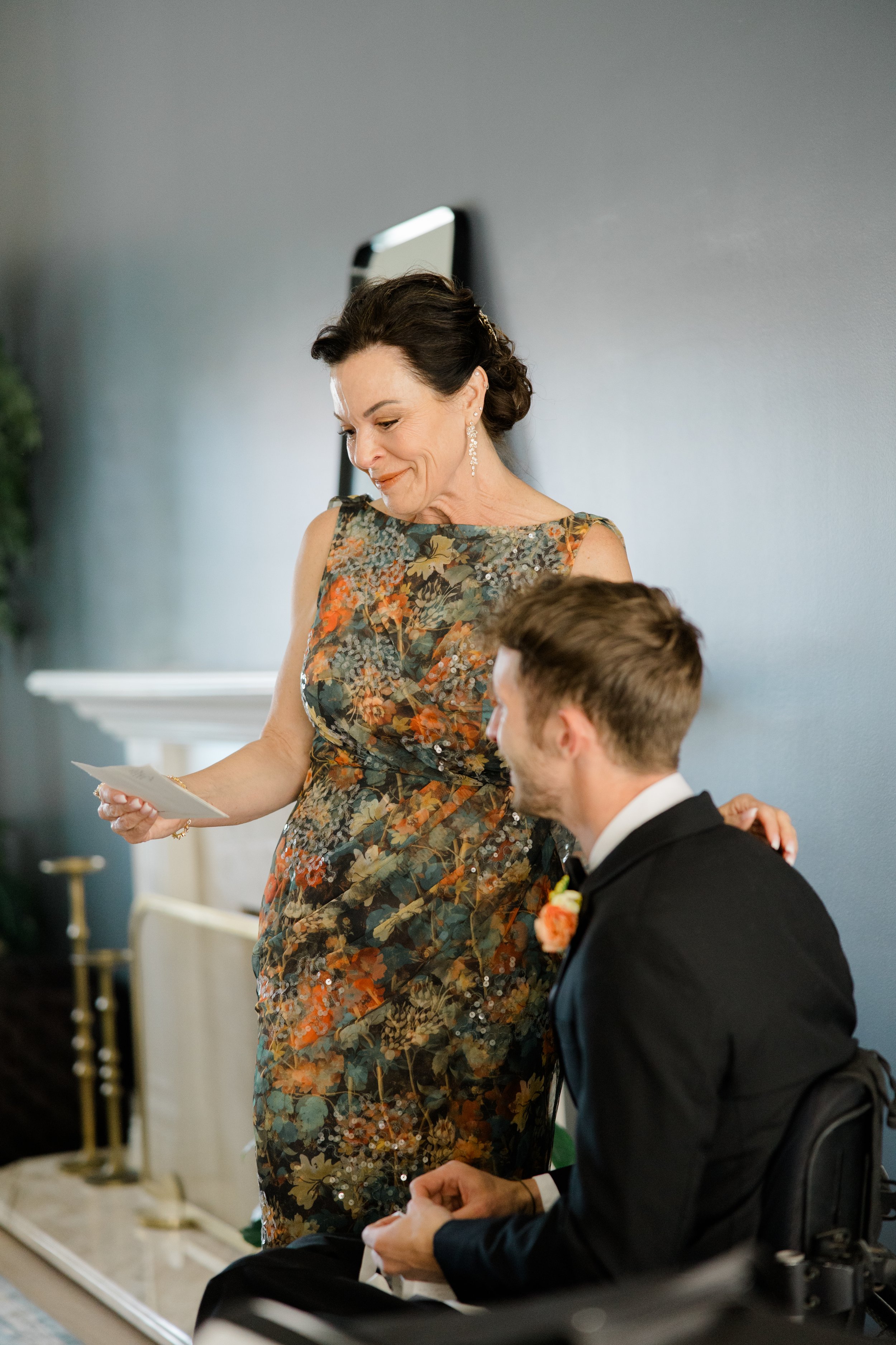 A woman in a floral dress holding a note, standing next to a man in a wheelchair dressed in a suit with a flower boutonniere, at a formal event inside a room with a gray wall and white fireplace