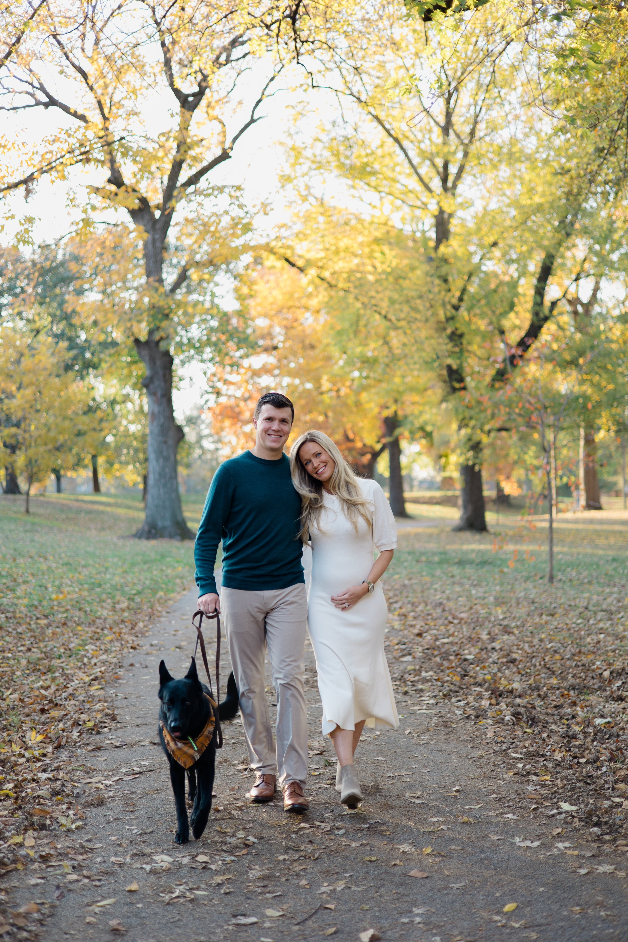 A smiling man and woman walking together on a leaf-covered path in a park during autumn, with a black dog wearing a bandana on a leash. The background features tall trees with yellow and green foliage and a bright sky.