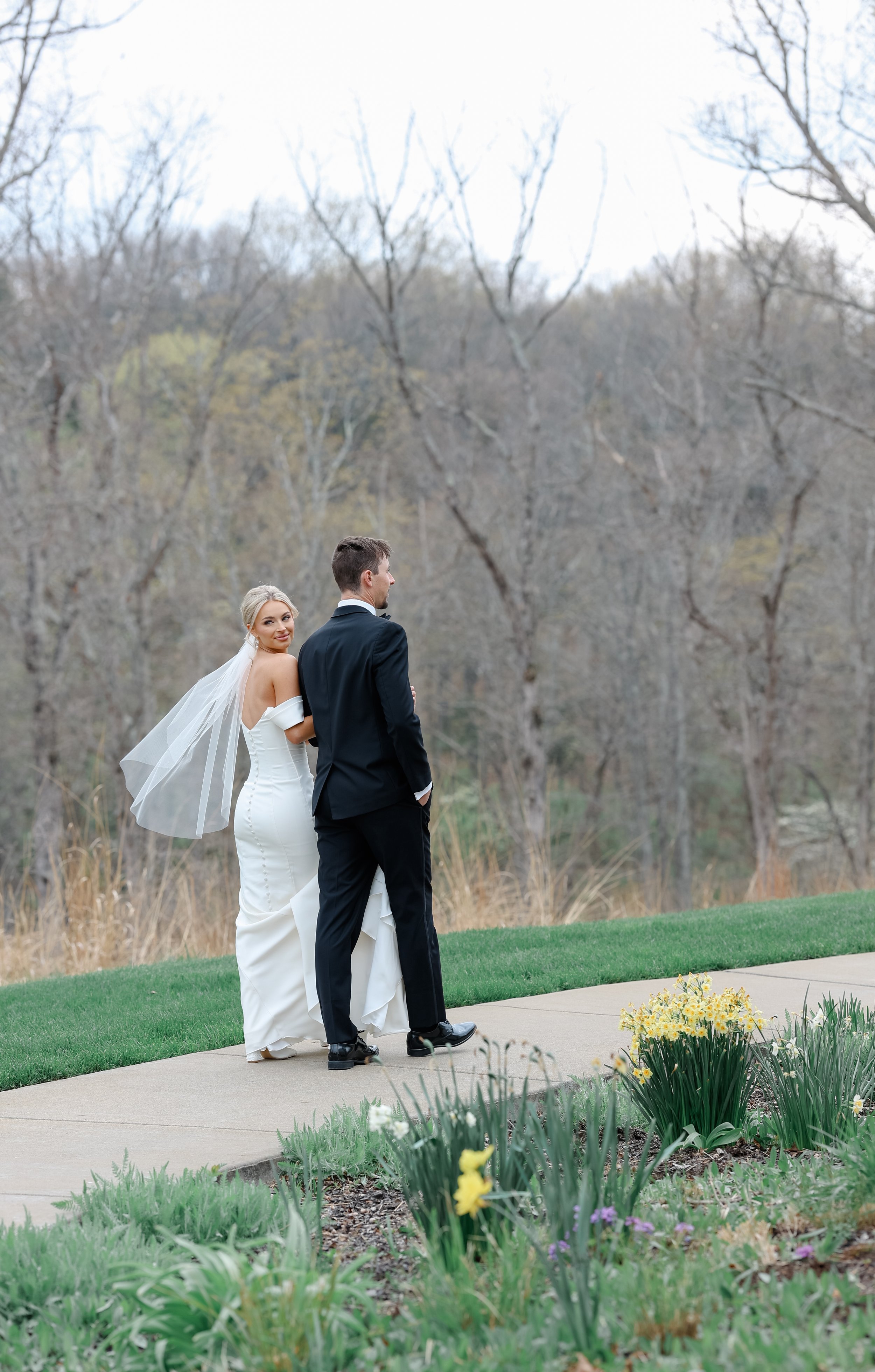 A bride and groom standing outdoors on a sidewalk, with trees and plants in the background, flowers on the ground, during a wedding ceremony.