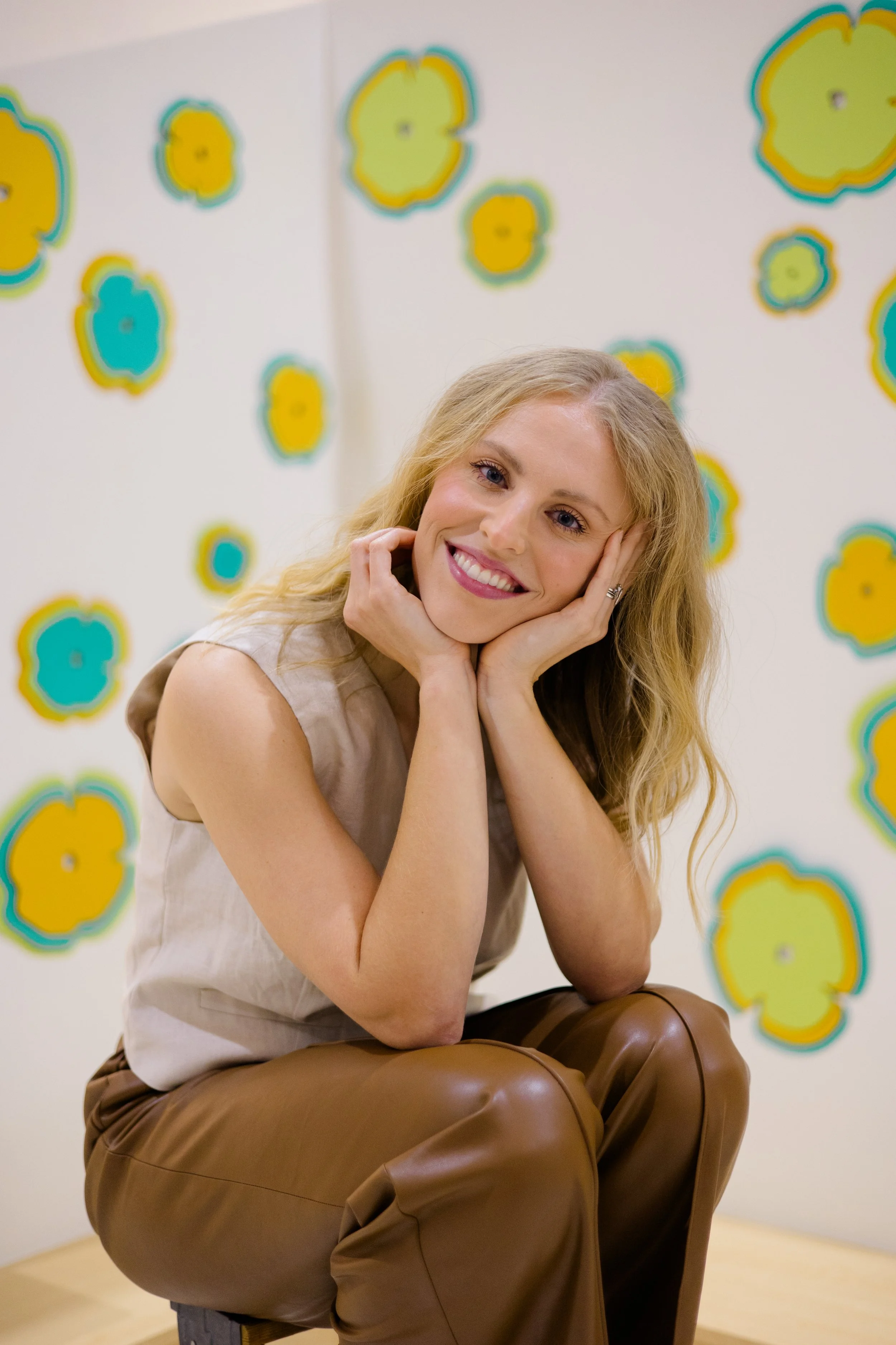 A woman with long blonde hair, smiling and resting her face on her hands, sitting on a chair with her knees up, in front of a white wall decorated with colorful, abstract floral patterns.