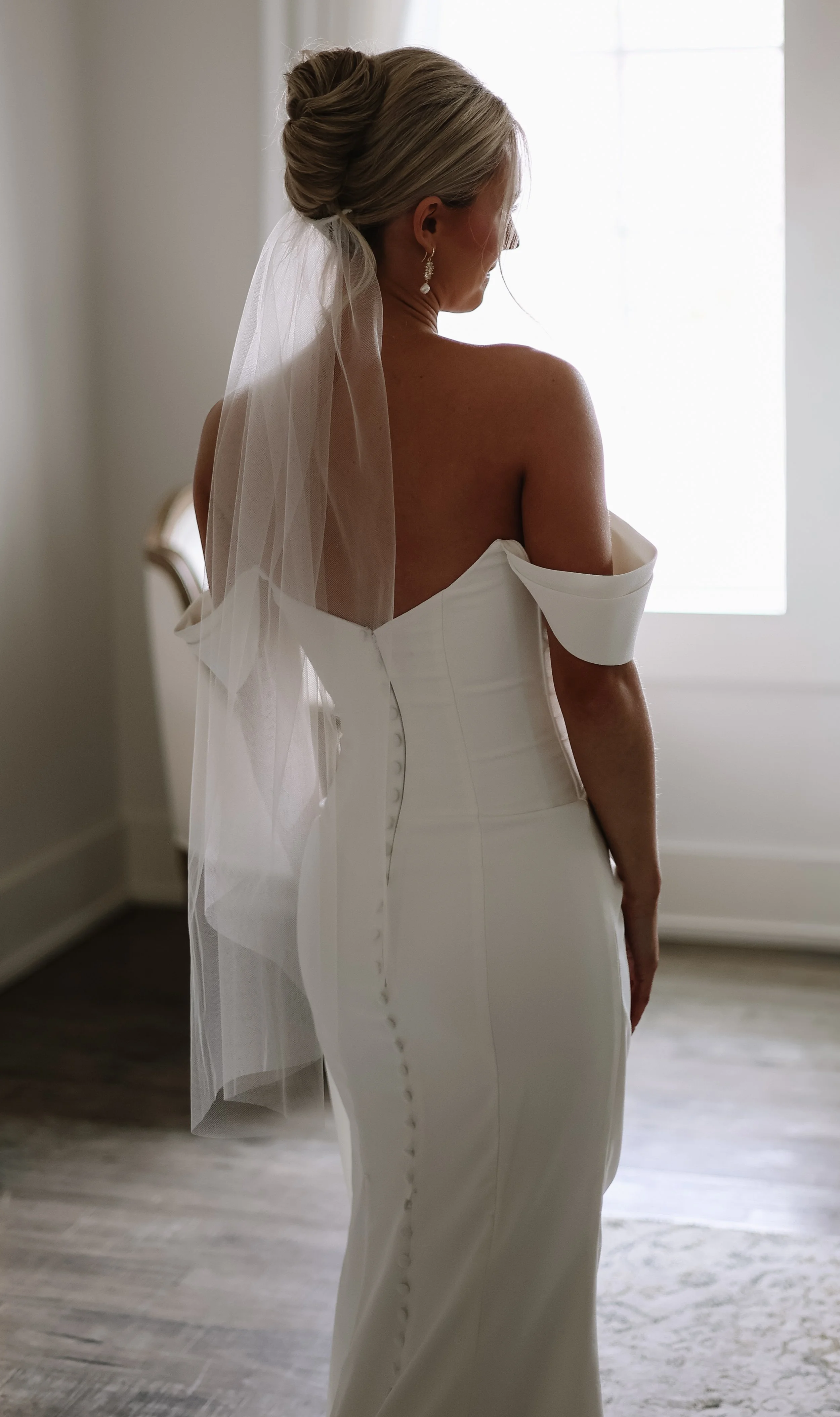 A bride in a white wedding dress with an off-the-shoulder design, standing indoors near a window with light streaming in. Her hair is styled in an elegant updo with a veil, and she is wearing pearl earrings. She is facing away from the camera, showca