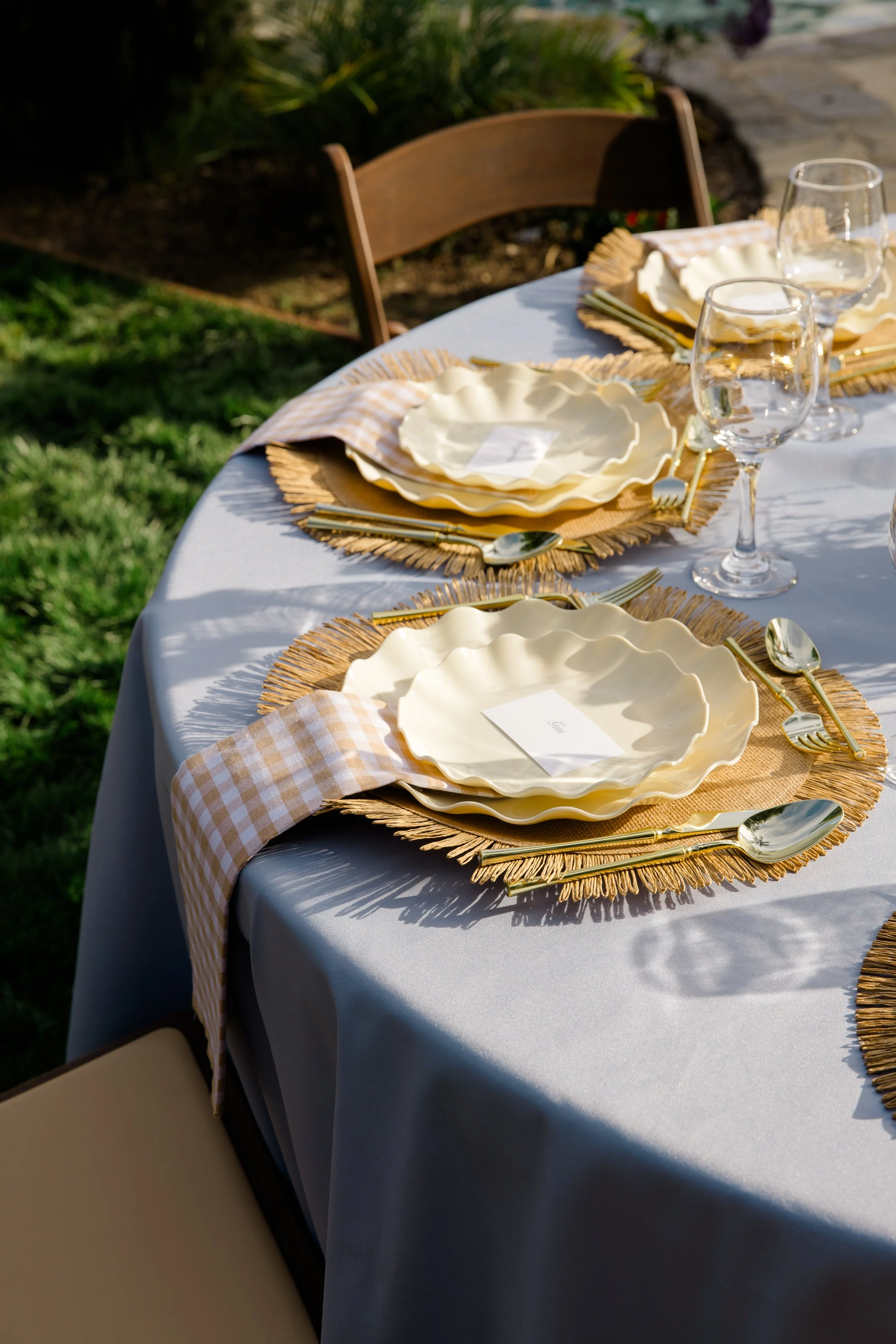 Outdoor dining table set with yellow and white checkered napkins, white plates with scalloped edges, gold utensils, clear wine glasses, and woven placemats, with a wooden chair and green plants in the background.