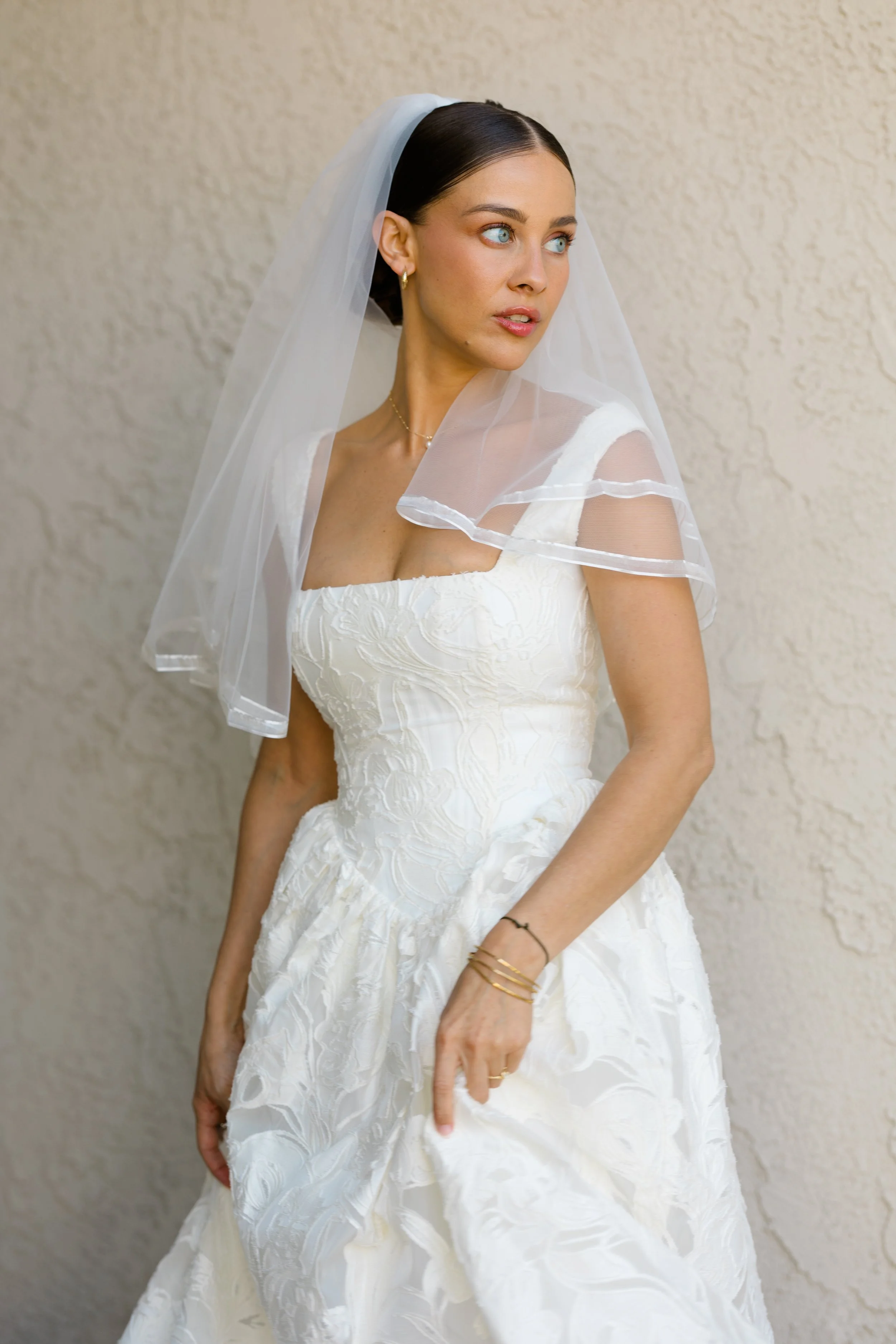 A woman in a white wedding dress with floral embroidery, short dark hair, blue eyes, wearing gold jewelry, and a sheer veil, standing against a beige textured wall.