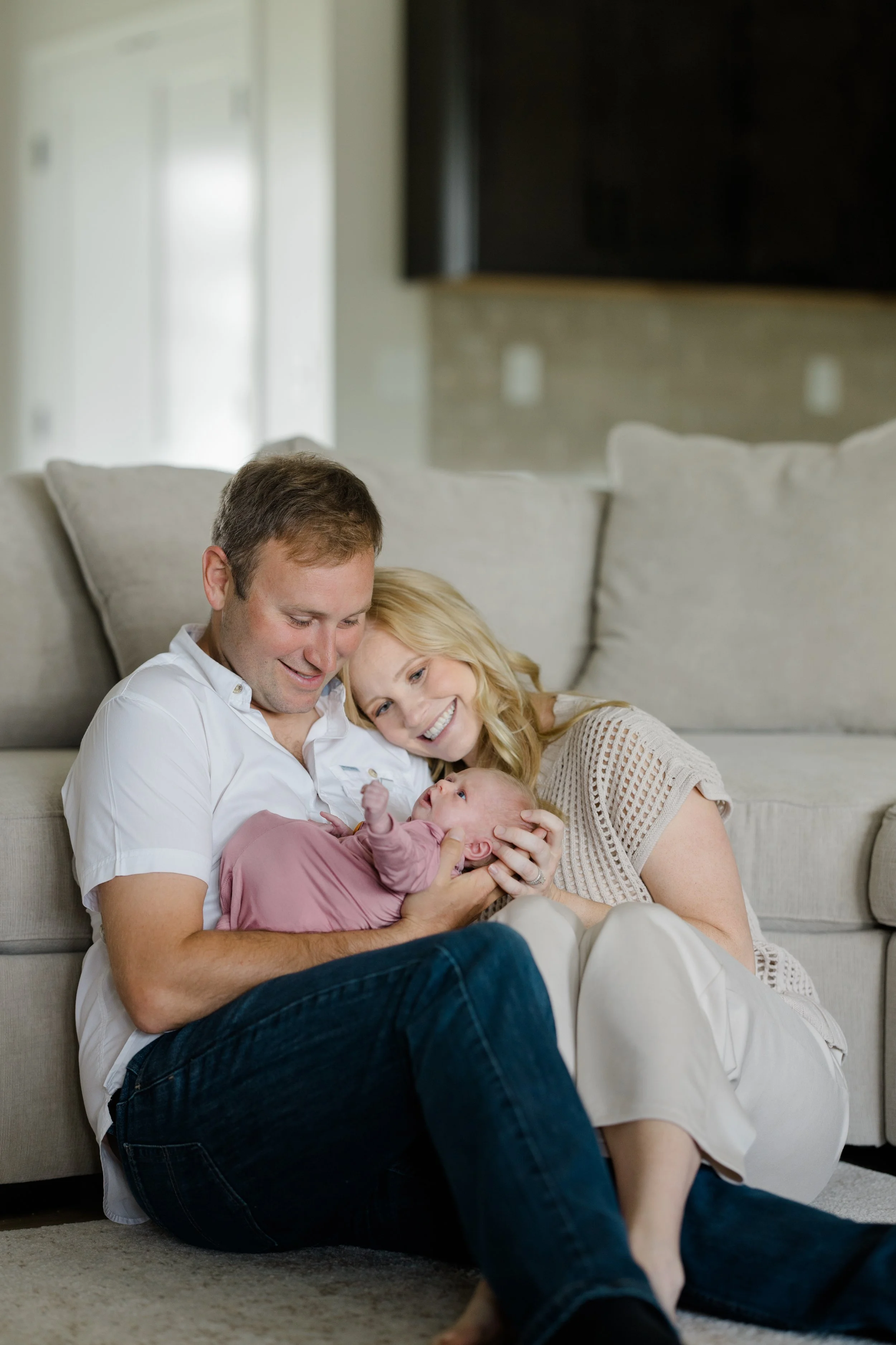A happy family sitting on the floor in front of a sofa, gathered closely together, smiling and looking at their baby.