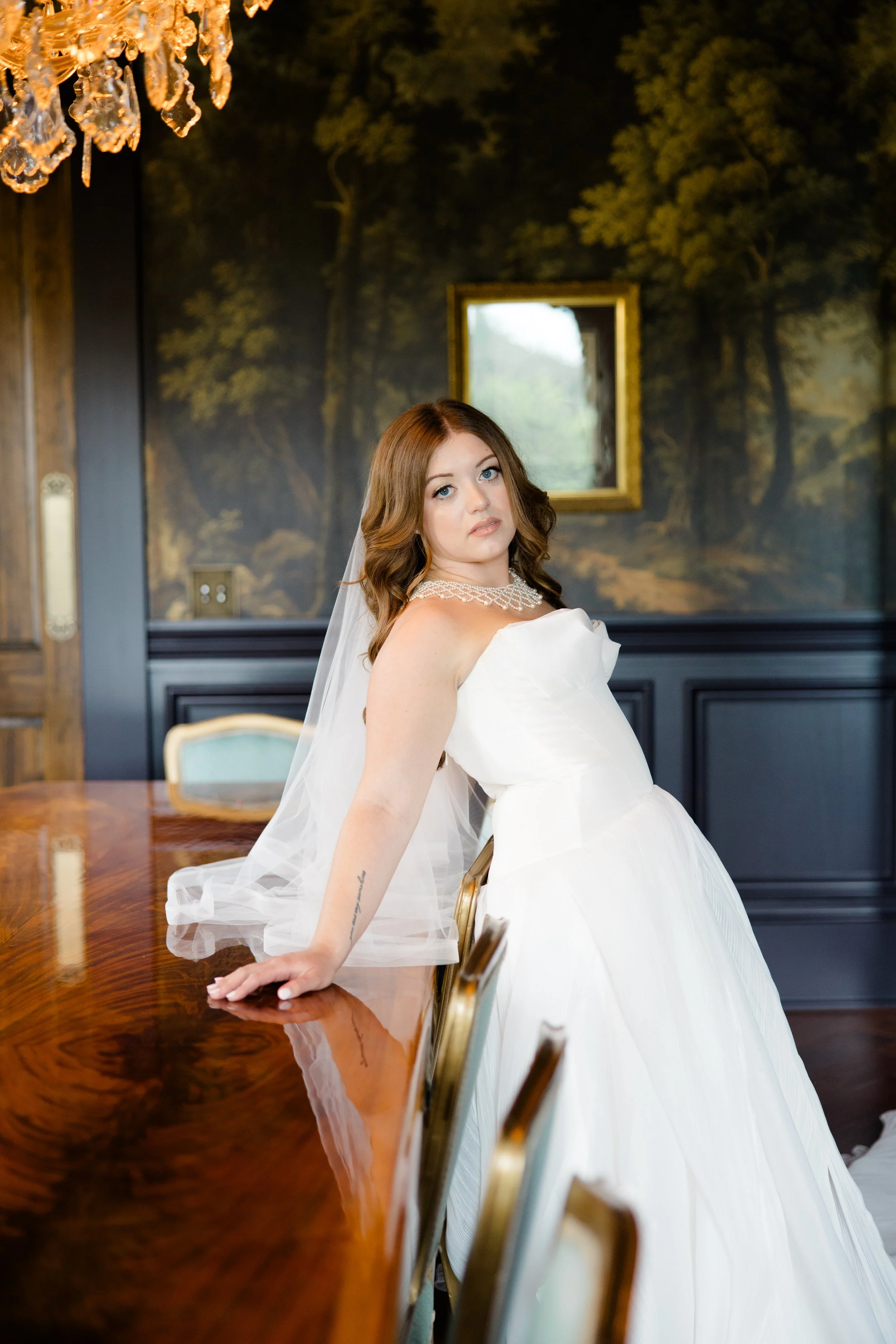 A woman in a white wedding dress with a pearl necklace, sitting on a chair in a room with dark blue wainscoting and a forest mural on the wall. She has long, wavy brown hair and is looking slightly to her left. There is a window with natural light be