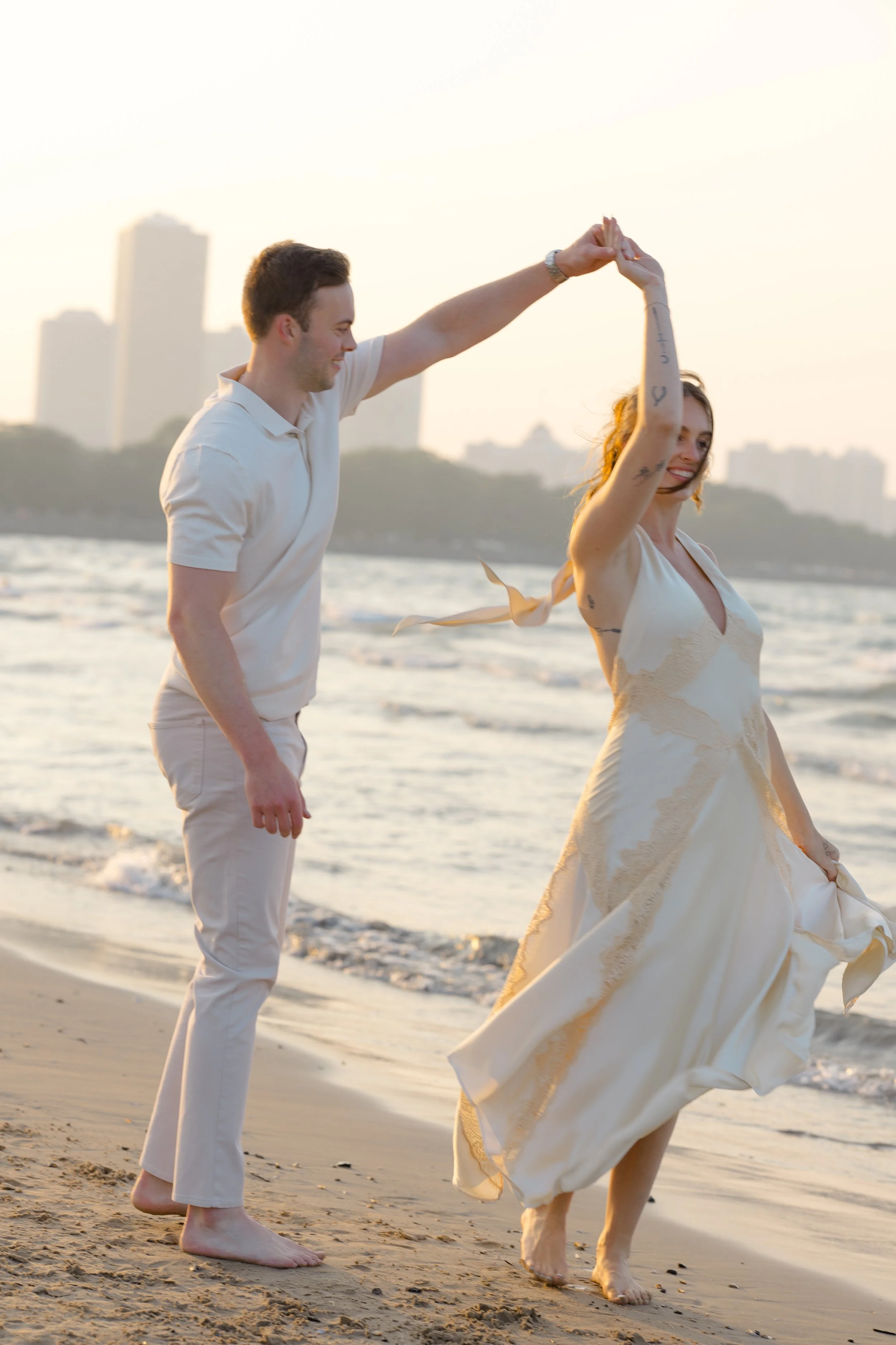 A couple dancing on the beach during sunset, with the city skyline in the background.