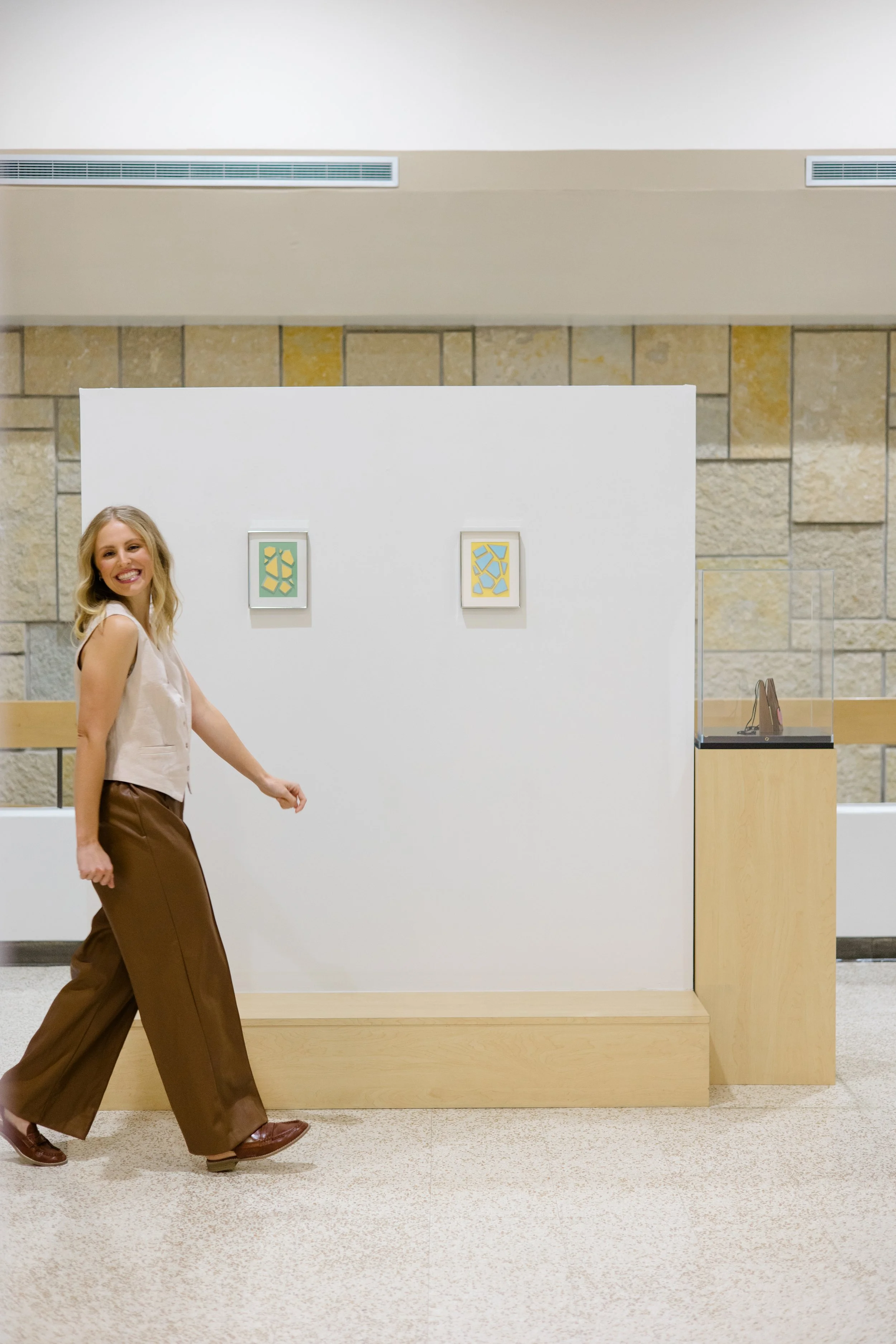 A woman with blonde hair smiling and walking past a display with framed artwork on a white wall in a gallery or museum.