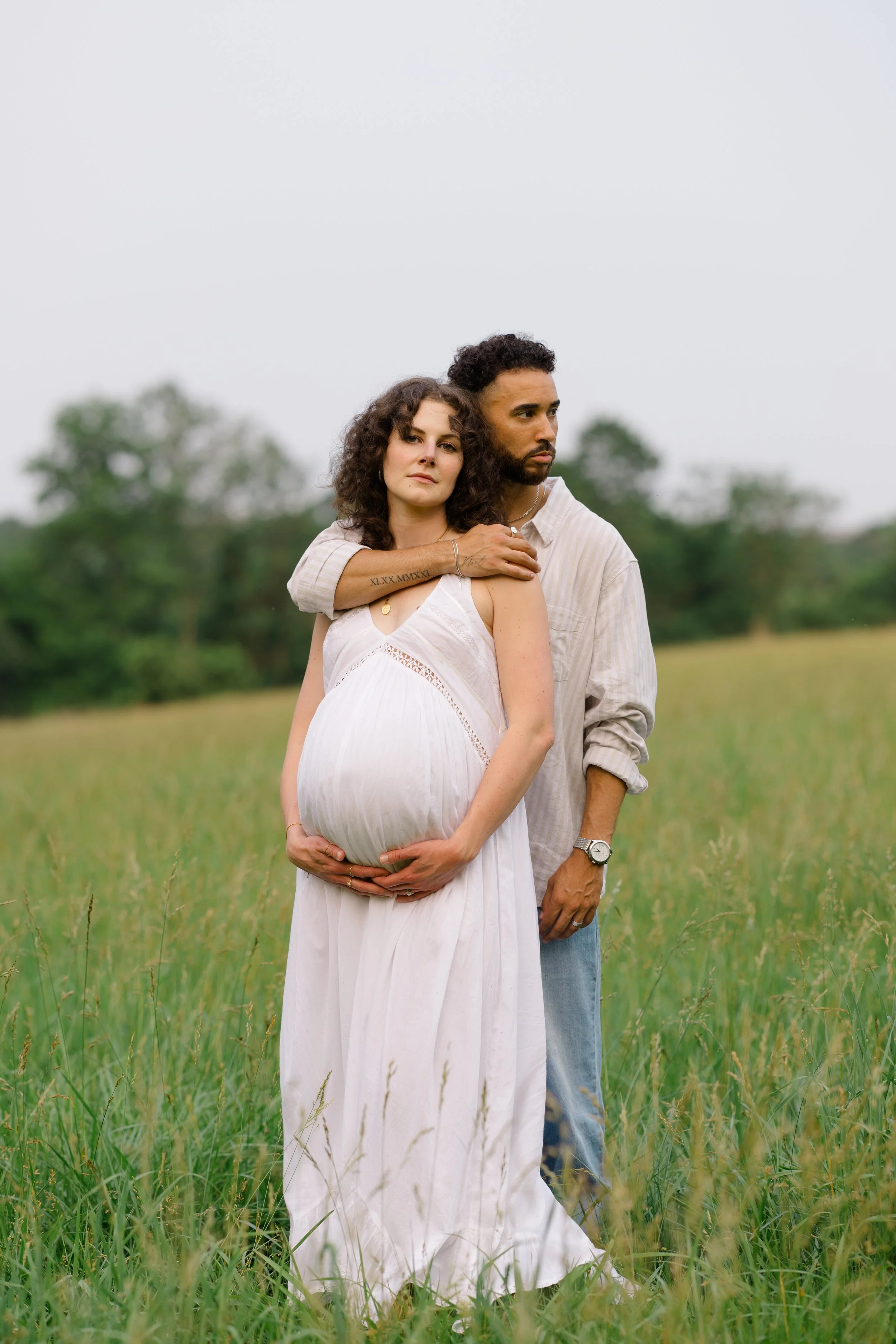 A pregnant woman in a white dress stands in a grassy field, holding her belly, while a man stands behind her with his arm around her shoulders, in an overcast outdoor setting with trees in the background.