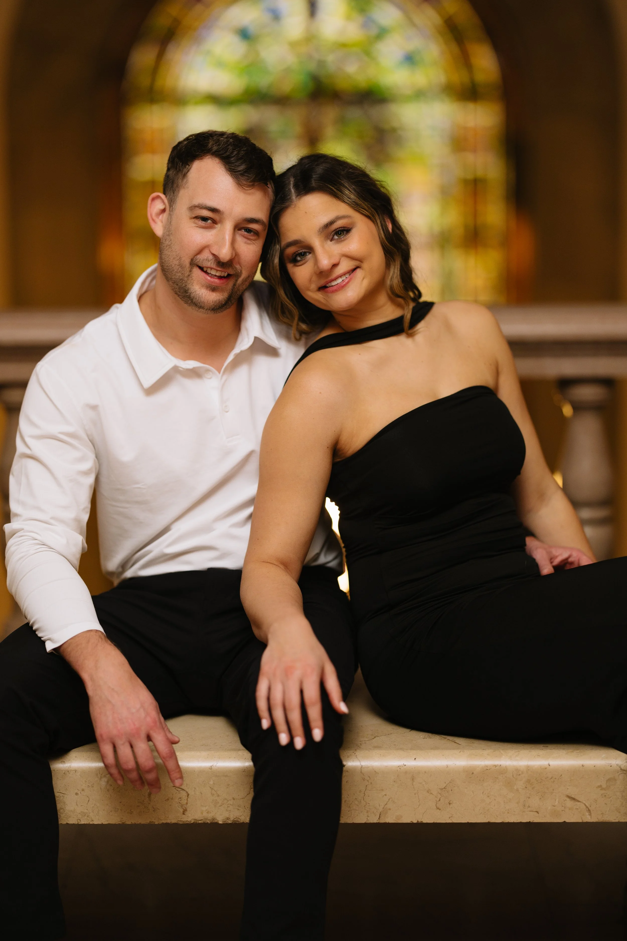 A happy couple sitting closely on a beige marble ledge inside a church or cathedral, with a stained glass window in the background. The man is wearing a white shirt and the woman is wearing a black strapless dress, both smiling at the camera.
