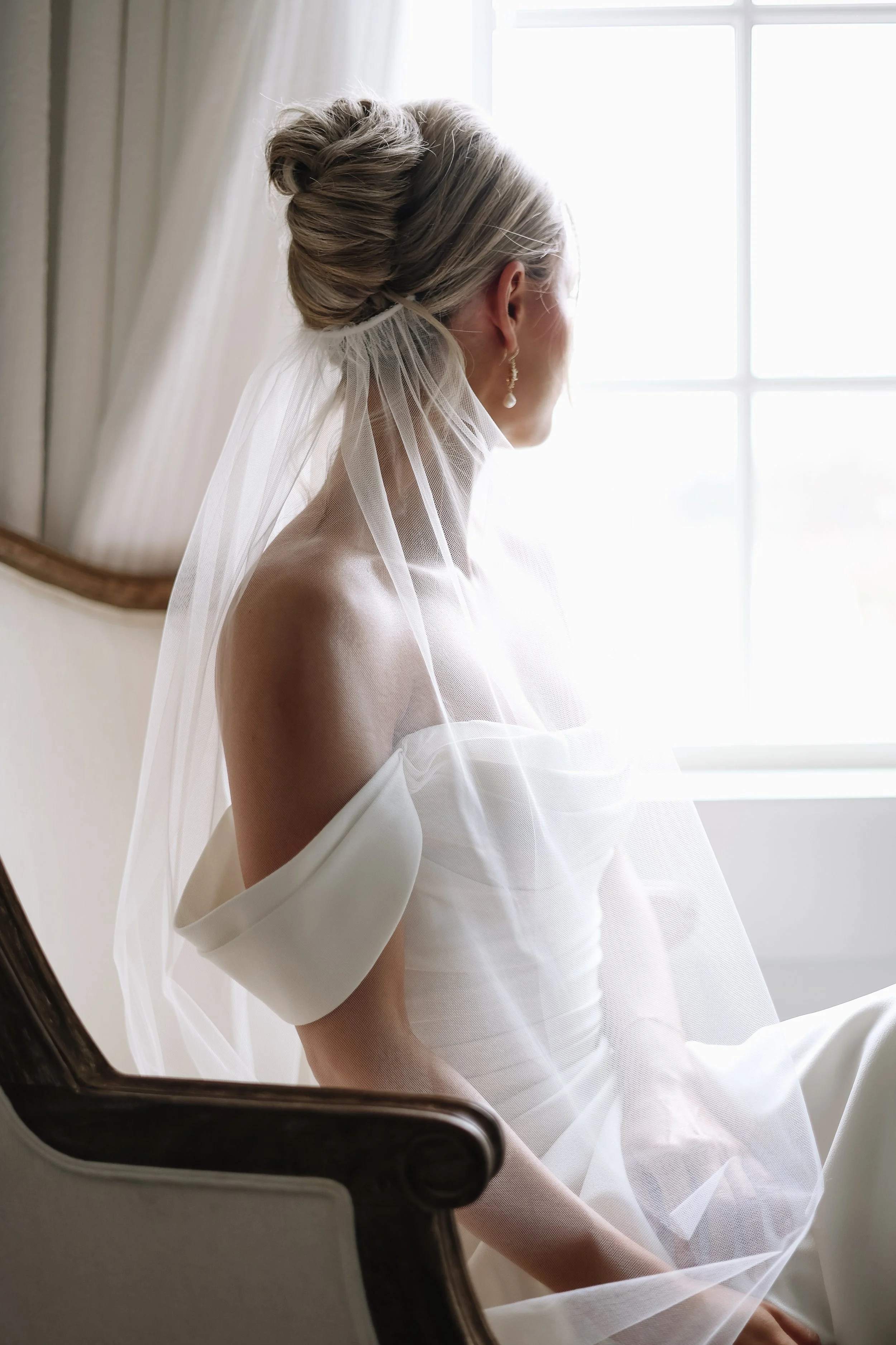 A woman with blonde hair styled in an updo, wearing a white strapless dress and pearl earrings, sitting by a window with sheer curtains, looking away from the camera.