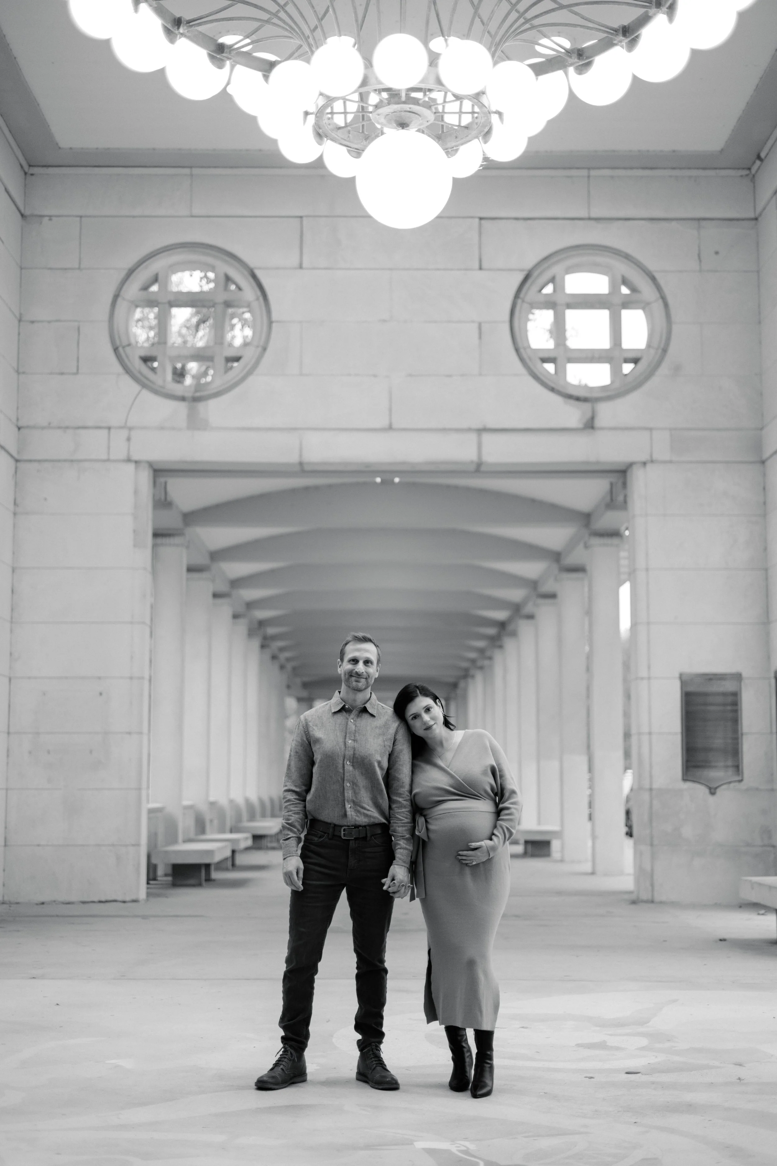 A black-and-white photo of a man and pregnant woman standing close together in a building corridor. The man has short hair, is wearing a button-up shirt, and dark pants. The woman has shoulder-length hair styled in loose waves, is wearing a long, fit
