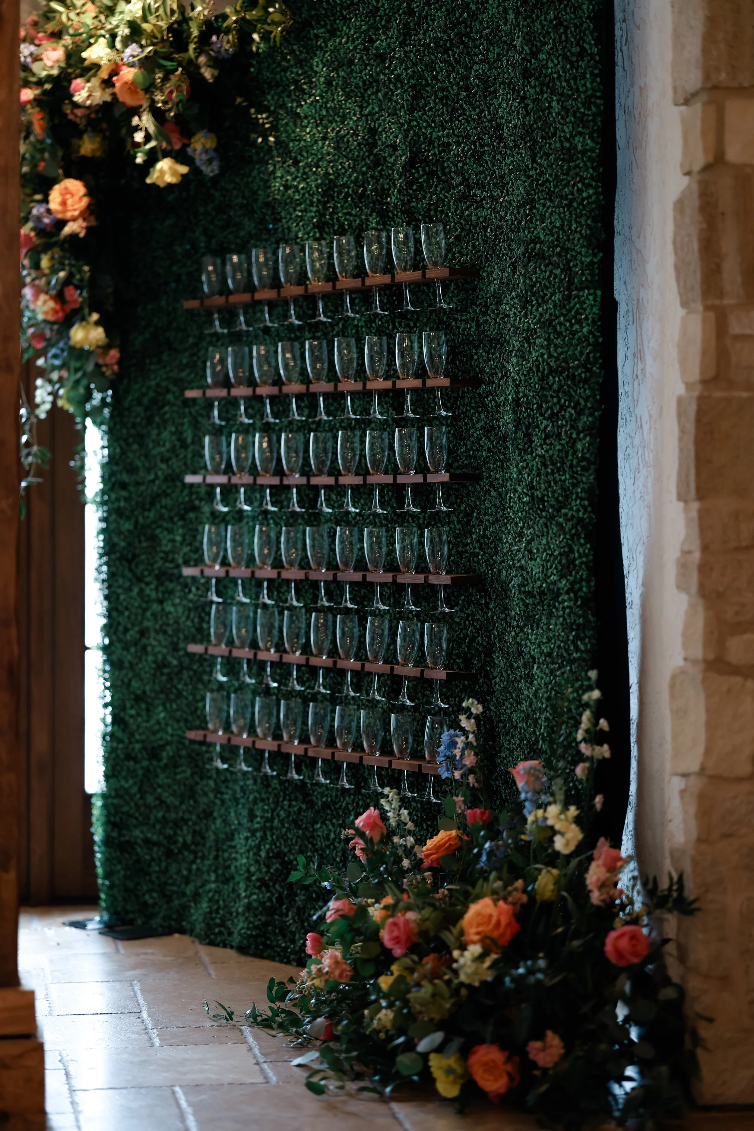 A decorated wall with lush green foliage, flowers, and multiple rows of empty champagne glasses on wooden shelves, with a tiled floor and a brick wall on the right.
