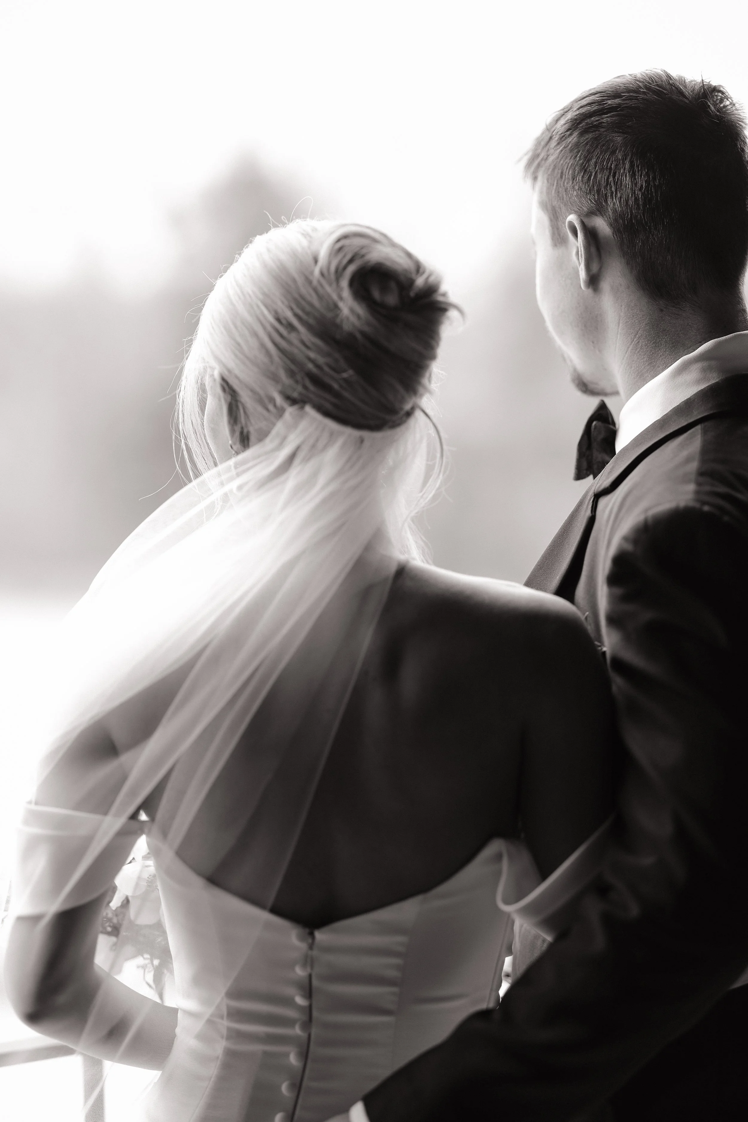 Black and white photo of a bride and groom from behind, sitting closely together, with the bride in a wedding dress and veil, and the groom in a tuxedo.
