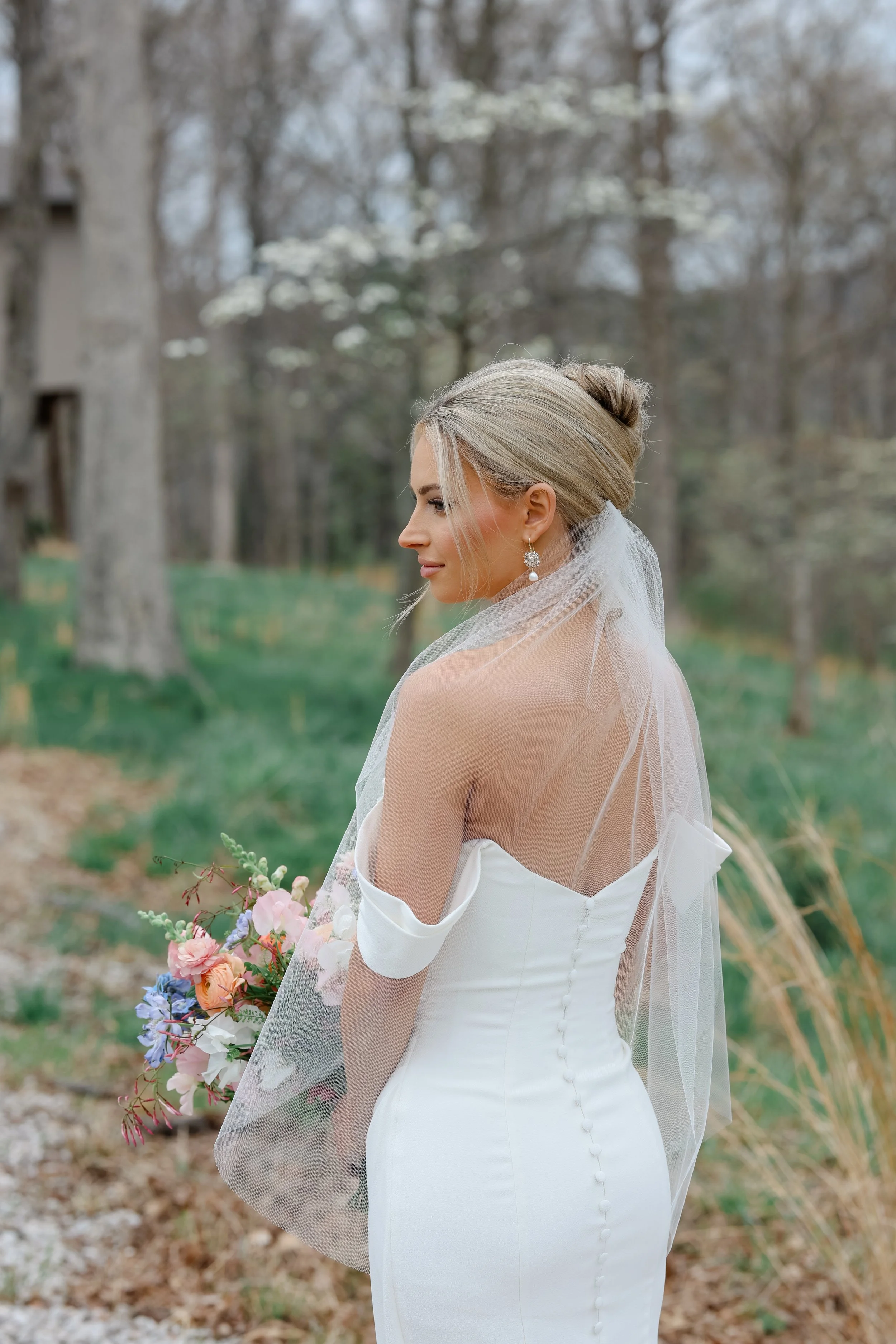 A bride with blonde hair styled in an elegant updo, wearing pearl earrings, a strapless white wedding dress with buttons on the back, holding a bouquet of pink, purple, and white flowers, standing outdoors in a wooded area with trees and greenery.