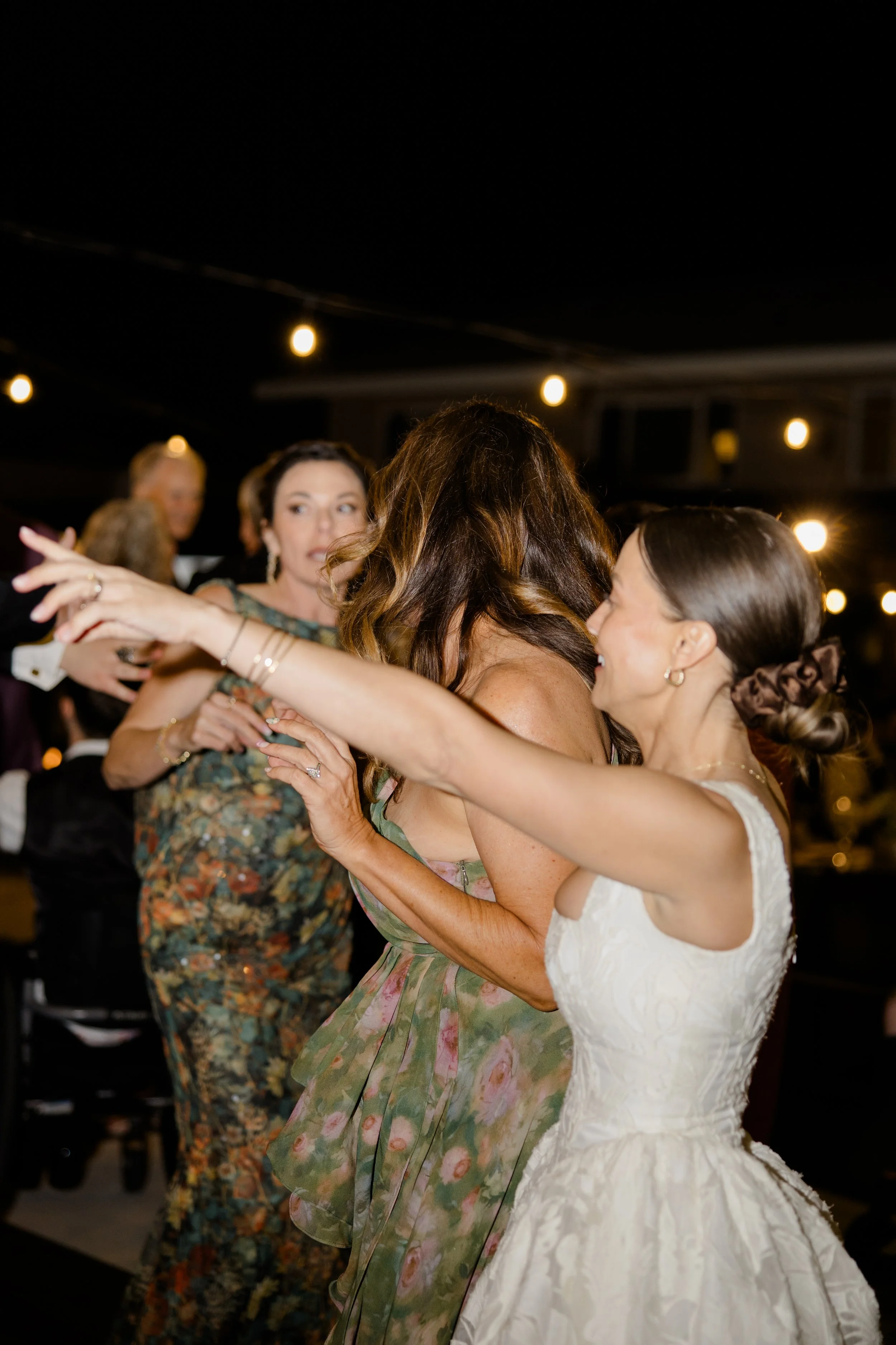 Two women dancing closely, with a woman in a white wedding dress and another in a colorful floral dress, at a celebration with hanging string lights in the background.