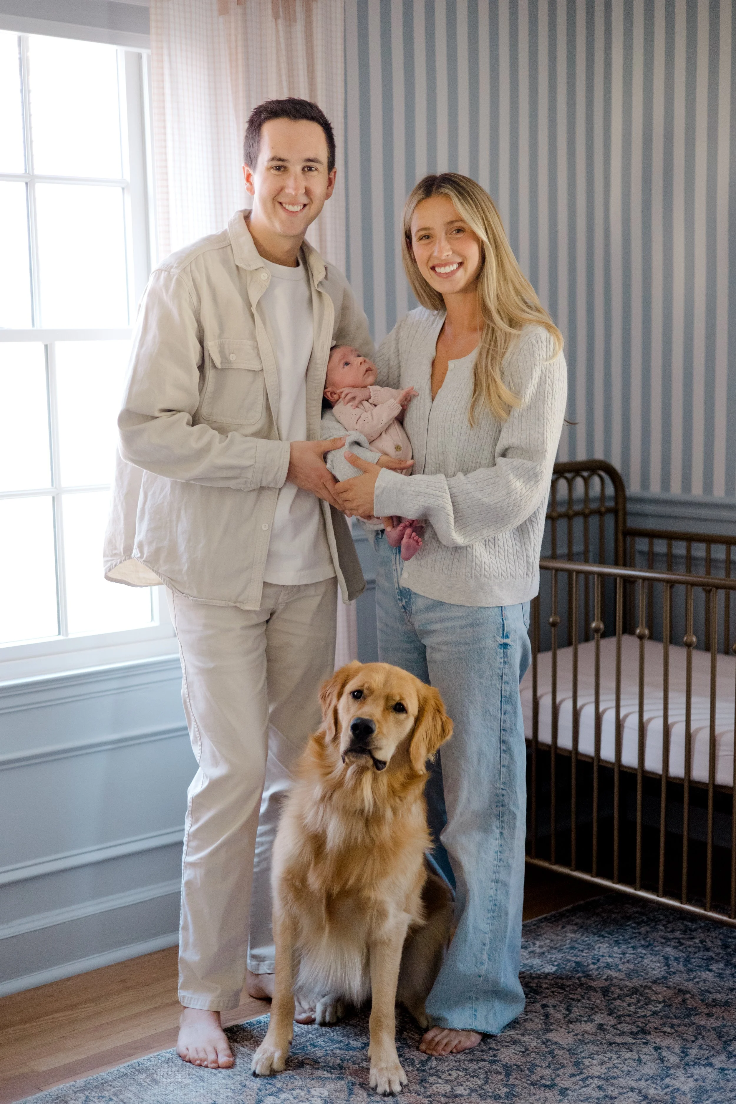 A happy couple with a newborn baby and a golden retriever dog inside a home. The man is holding the baby, and the woman is standing next to them, all smiling. The dog is sitting on the carpet in front of them.