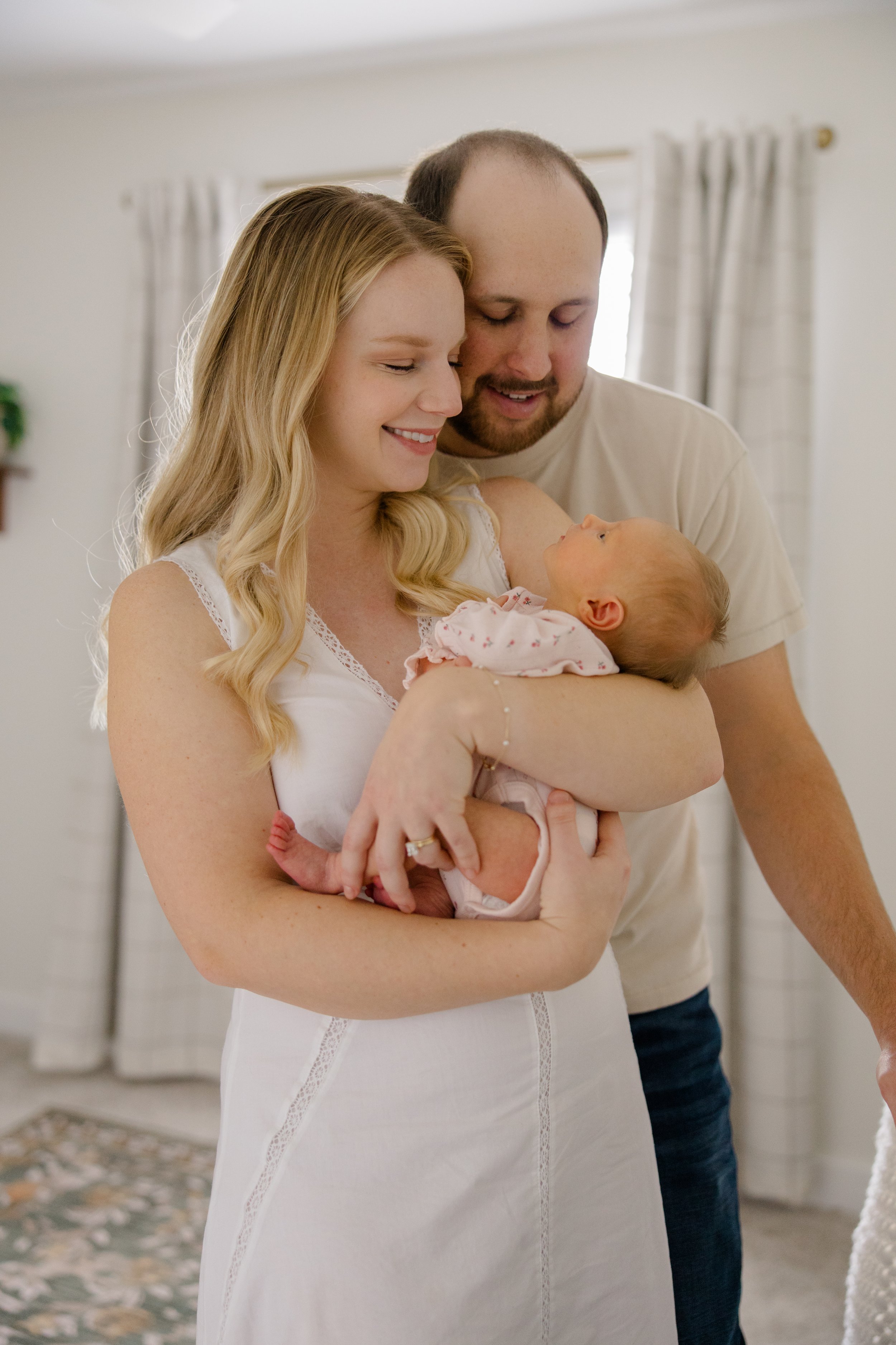 A couple holding a newborn baby in a brightly lit room with white walls, curtains, and a patterned rug.