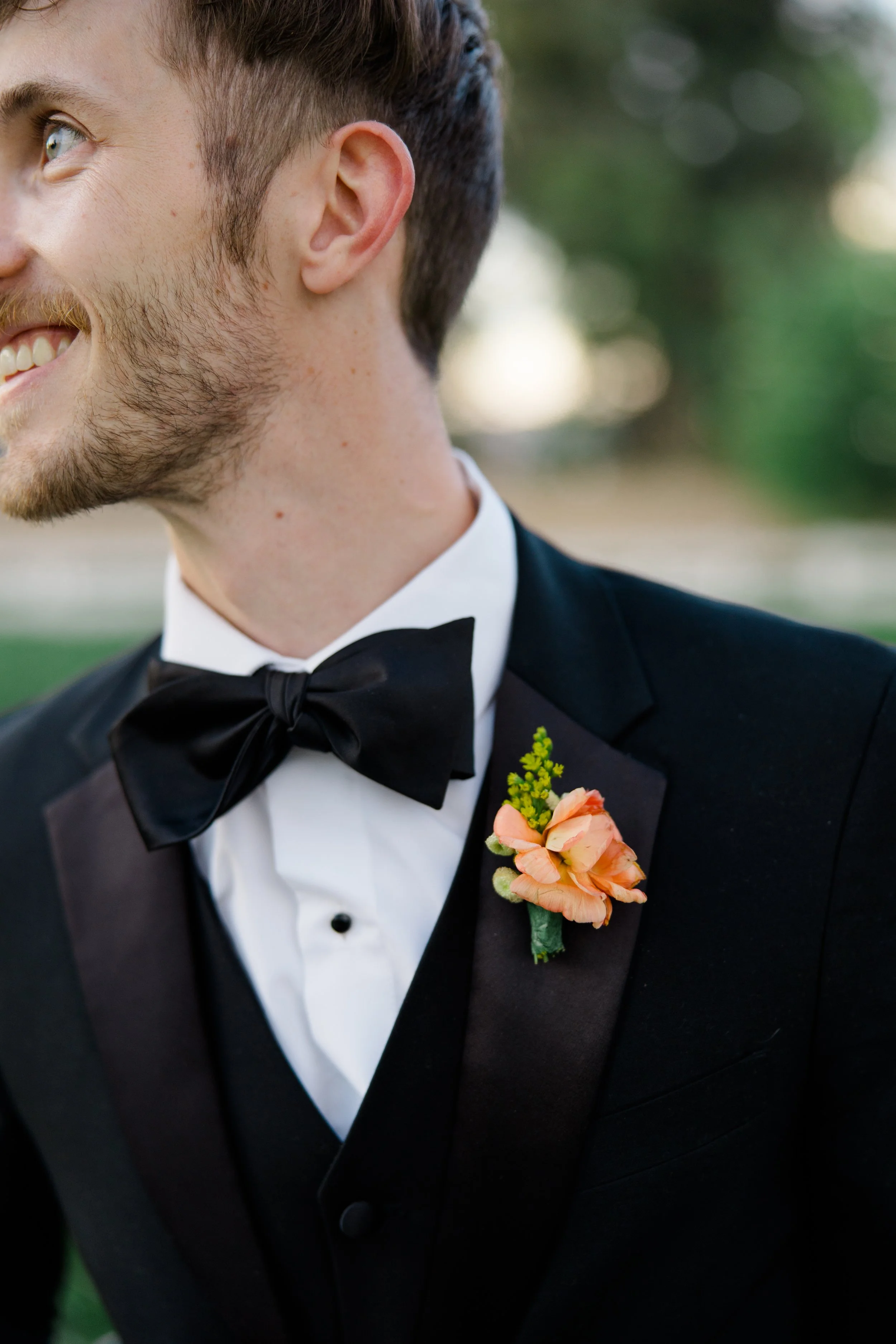 Close-up of a man in a tuxedo with a bow tie and a boutonniere, smiling outdoors.