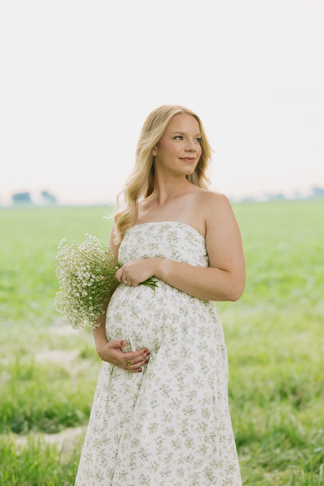 A pregnant woman holding a bouquet of white flowers outdoors in a green field during daytime, looking slightly to her left.