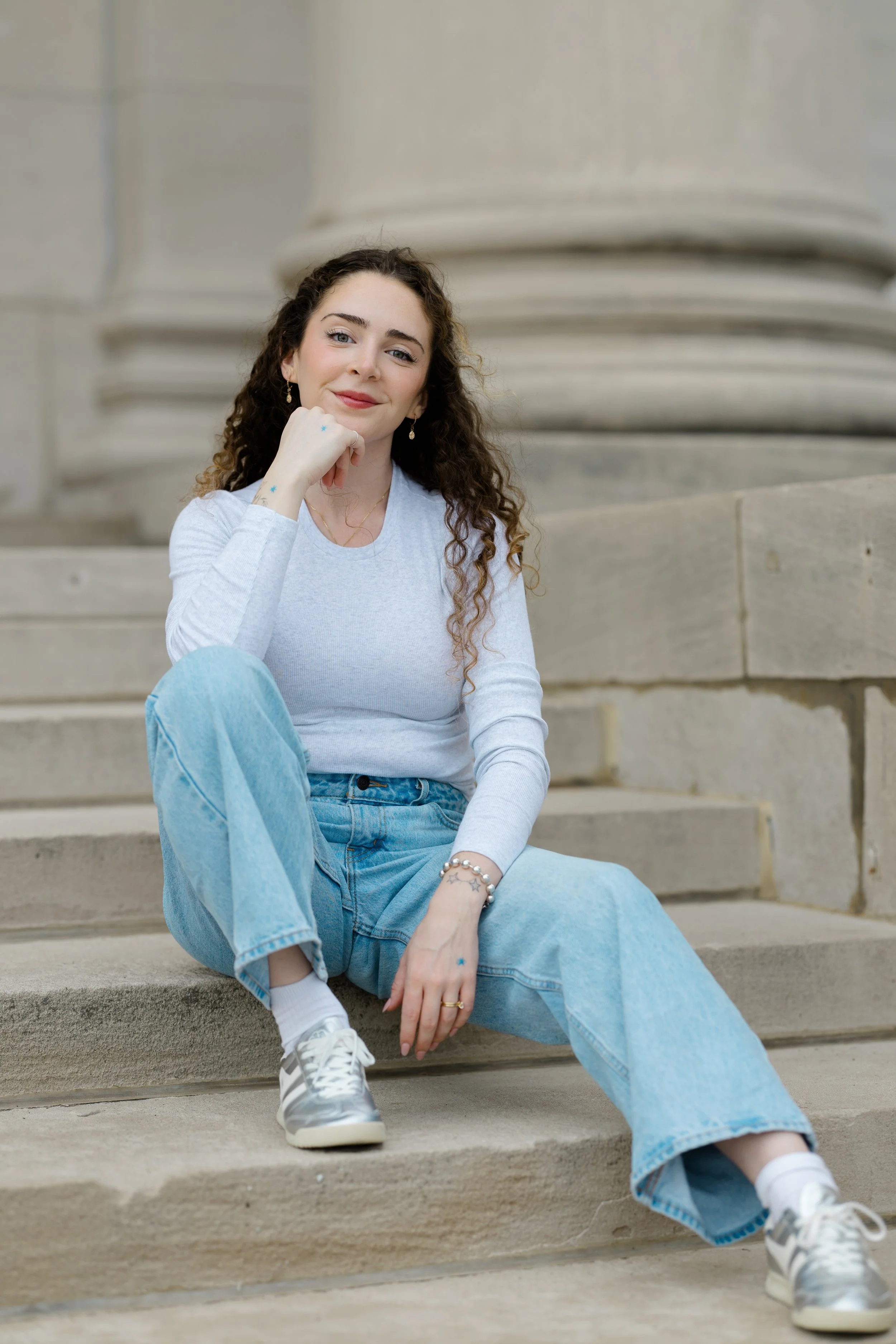 A young woman with long, curly brown hair sitting on outdoor stone steps, wearing a light gray long-sleeve shirt, light blue jeans, white sneakers, and jewelry, smiling and resting her chin on her hand.