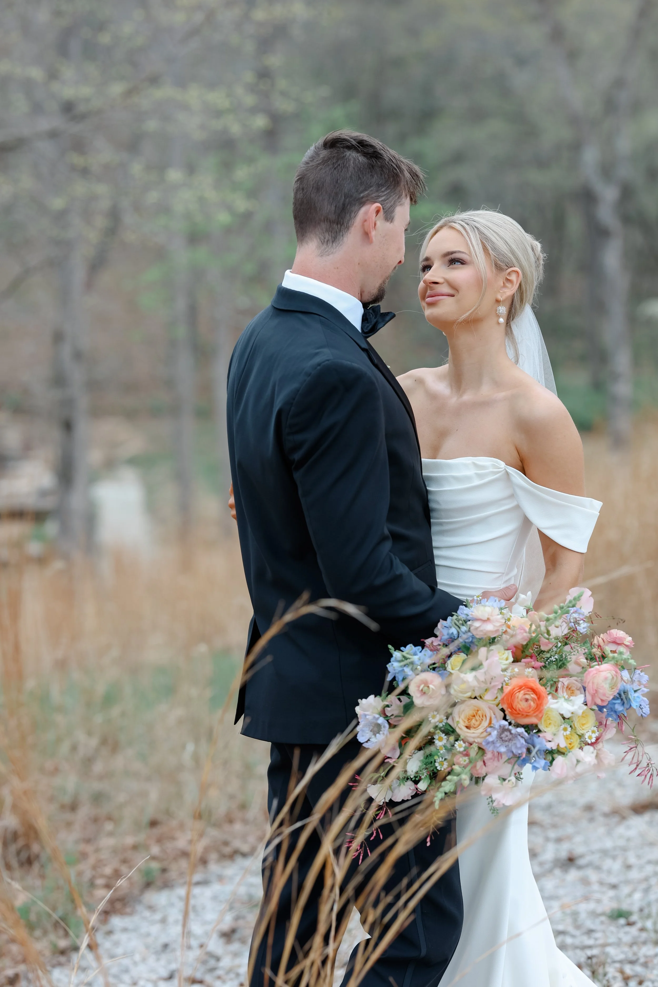 A bride and groom stand close together outdoors, facing each other, with the bride holding a large bouquet of colorful flowers. The bride has blonde hair and is wearing a strapless white wedding gown and pearl earrings. The groom has dark hair and is