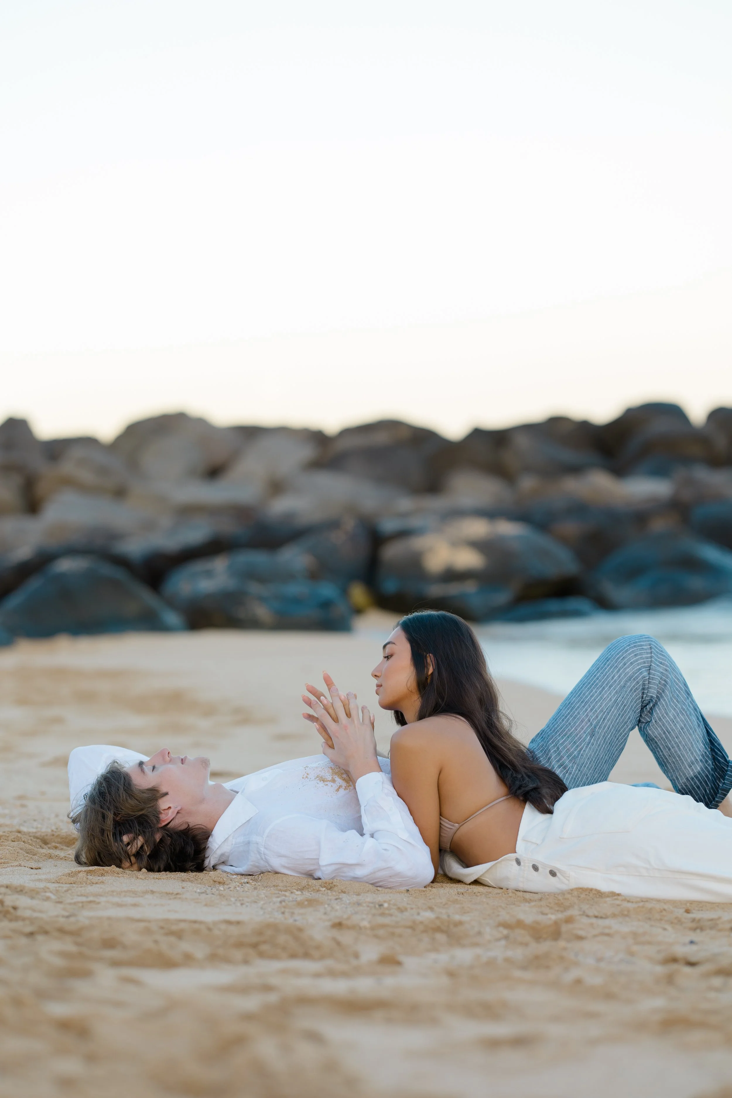 Two women are lying on a sandy beach facing each other, with large rocks in the background. One woman has dark hair and is sitting up, while the other woman has light brown hair and is lying down.