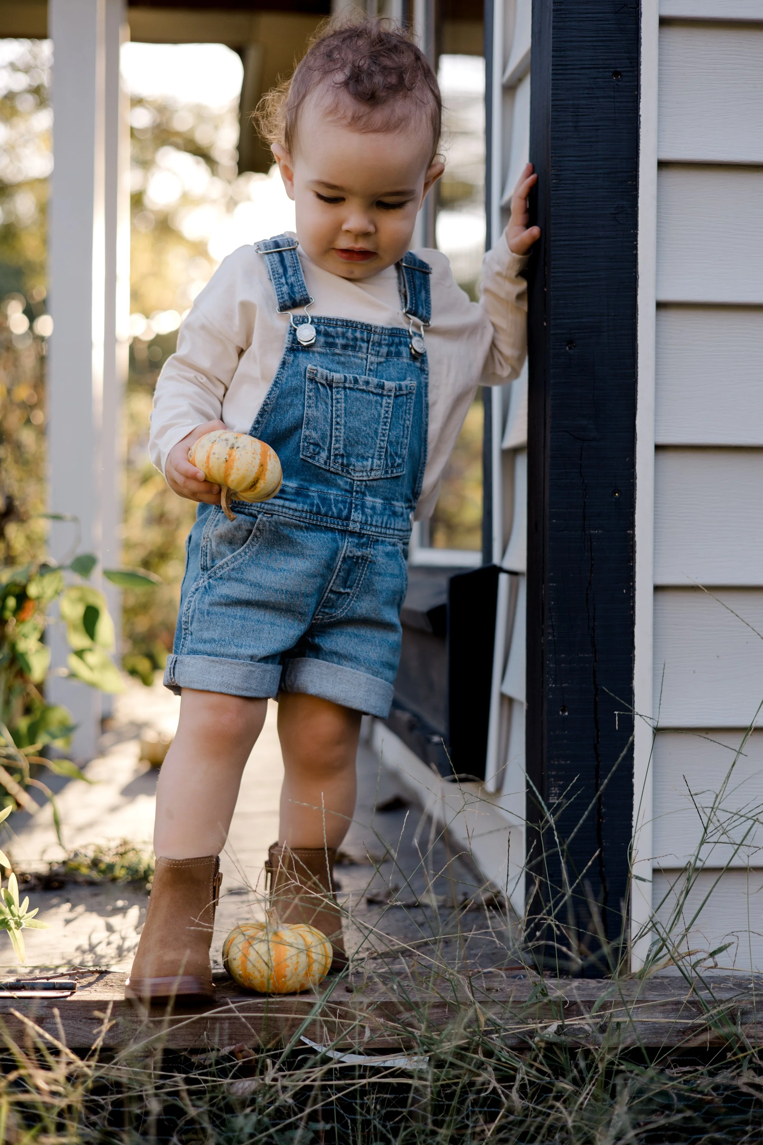 A young child in denim overalls and brown boots holding a small pumpkin, standing outside near a house with sunset lighting.