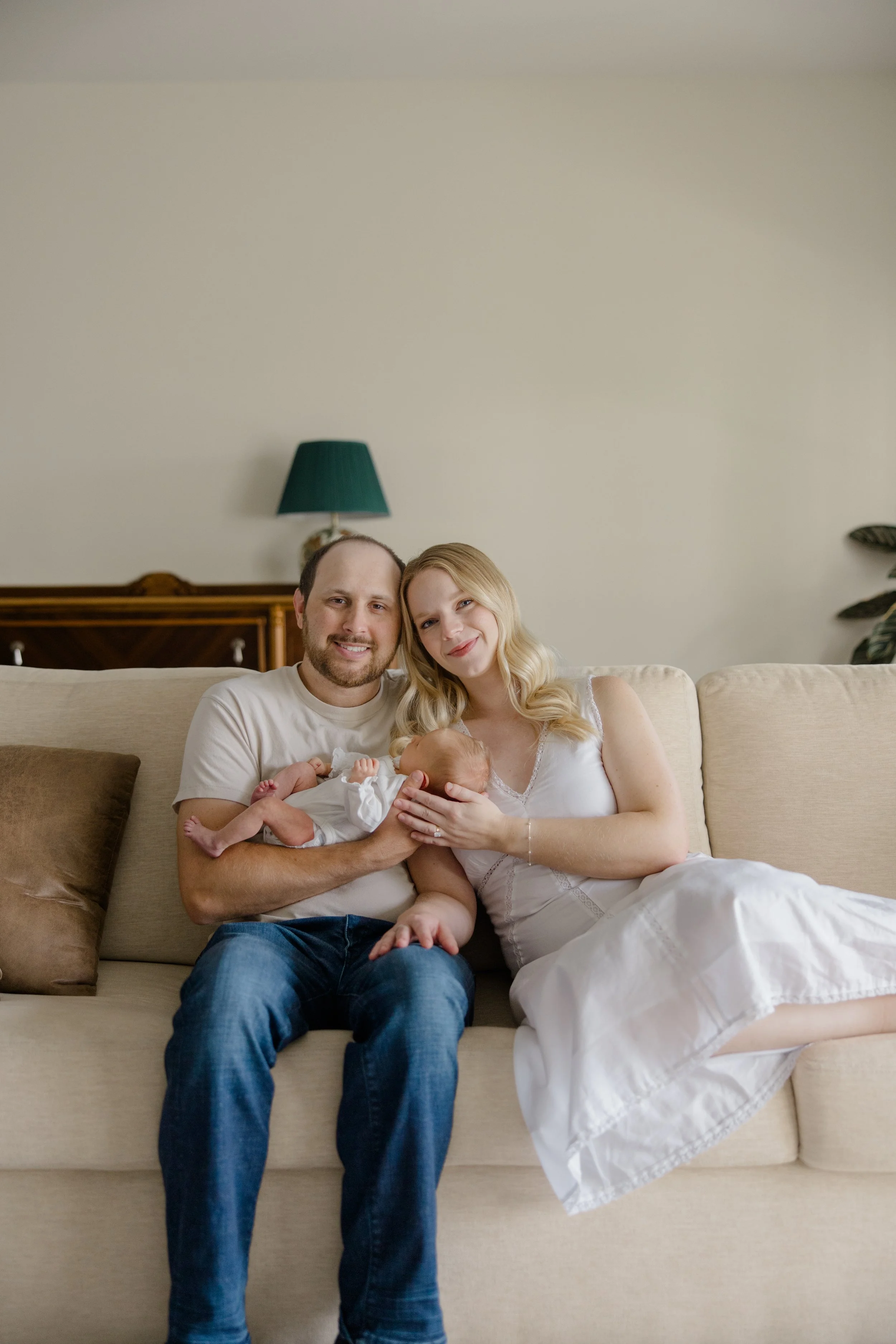 A family sitting on a beige couch in a living room, holding a newborn baby, with a woman and a man smiling at the camera.