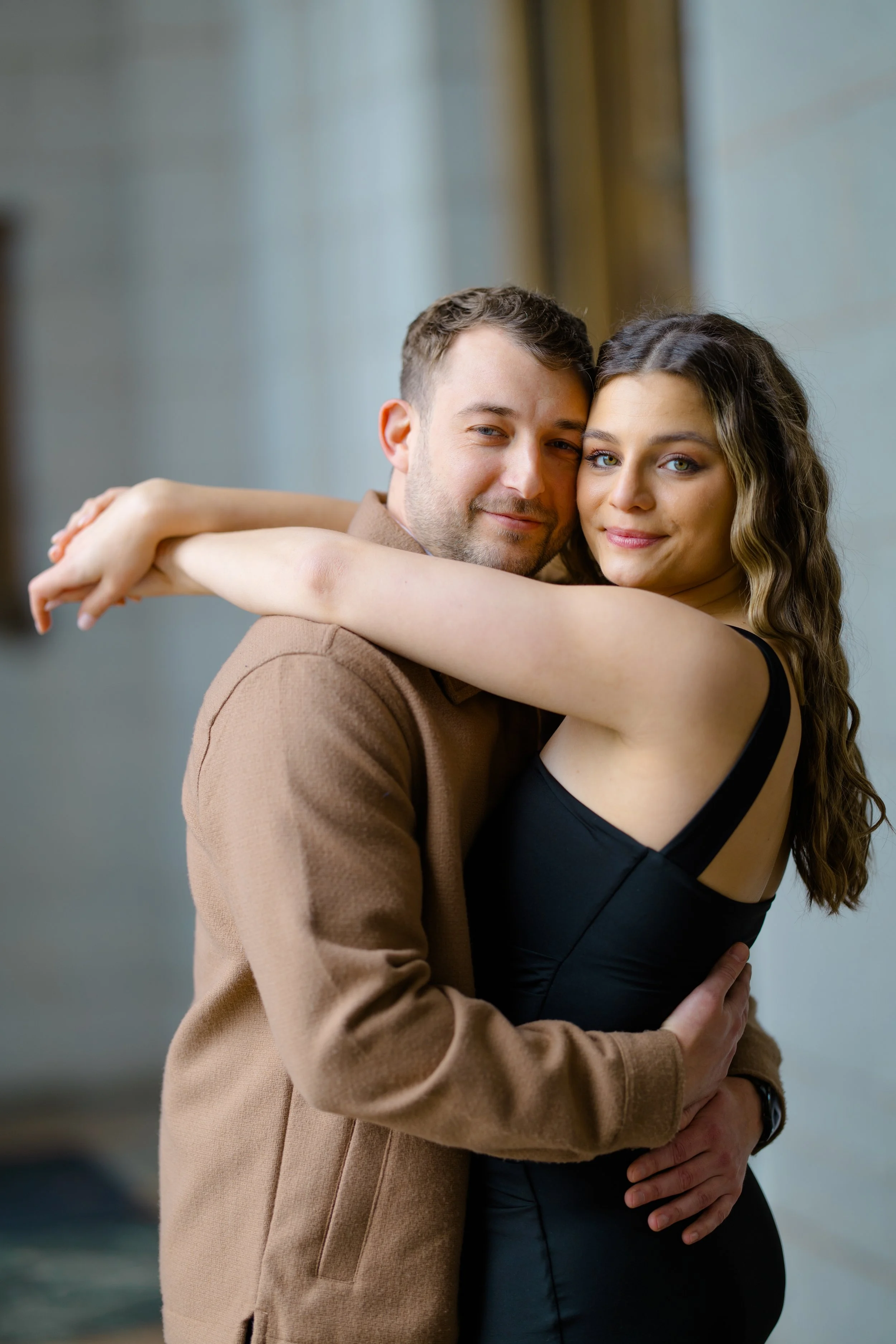 A couple hugging, with the man wearing a brown blazer and the woman in a black dress, standing indoors with blurred background.