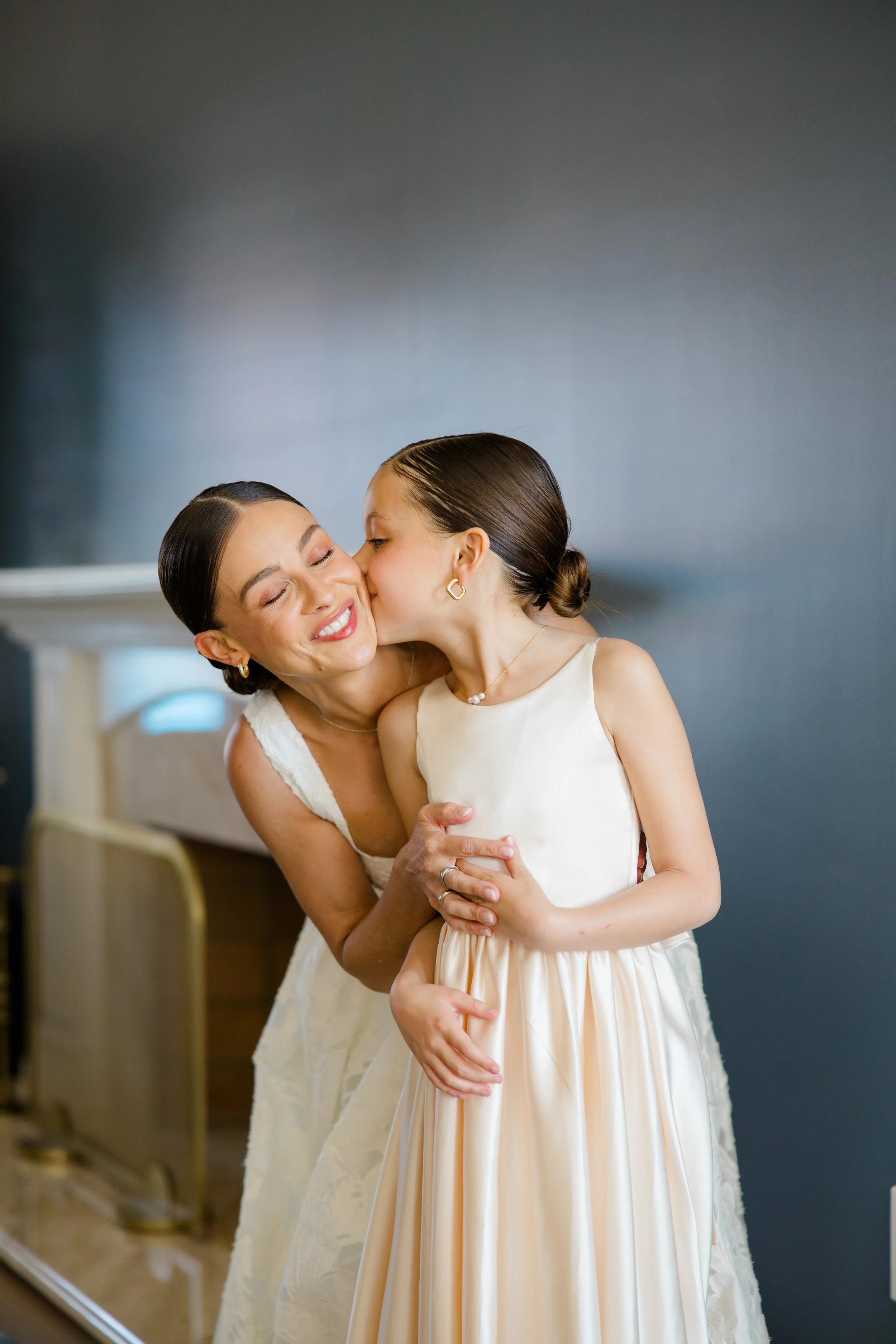 Two women, one older and one younger, sharing a kiss on the cheek in a room with a blue wall and a mirror in the background. They are both dressed in elegant, light-colored dresses and have their hair neatly styled.