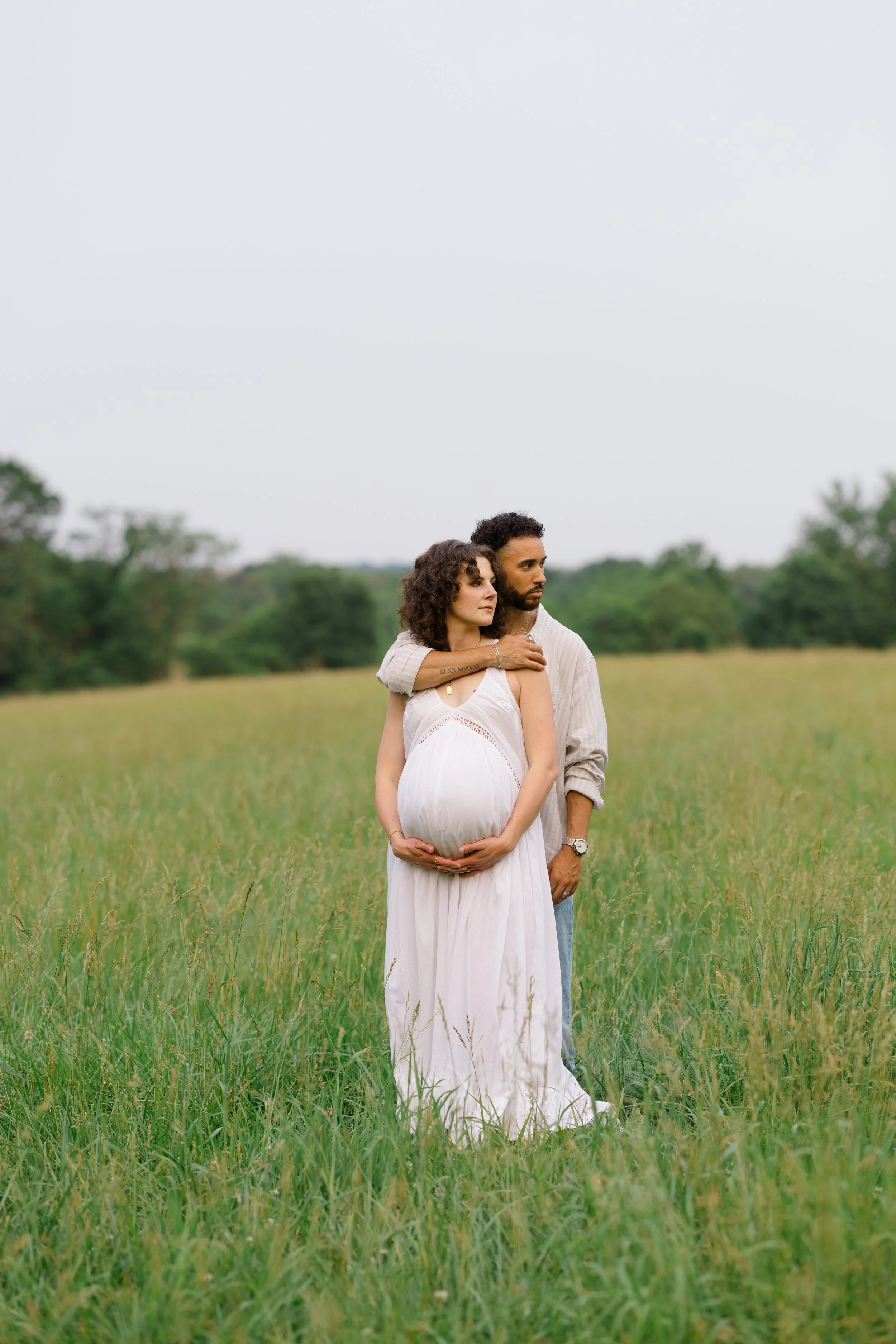 A pregnant woman and a man standing in a grassy field, with the woman embracing her belly and the man standing behind her, both looking to the side.