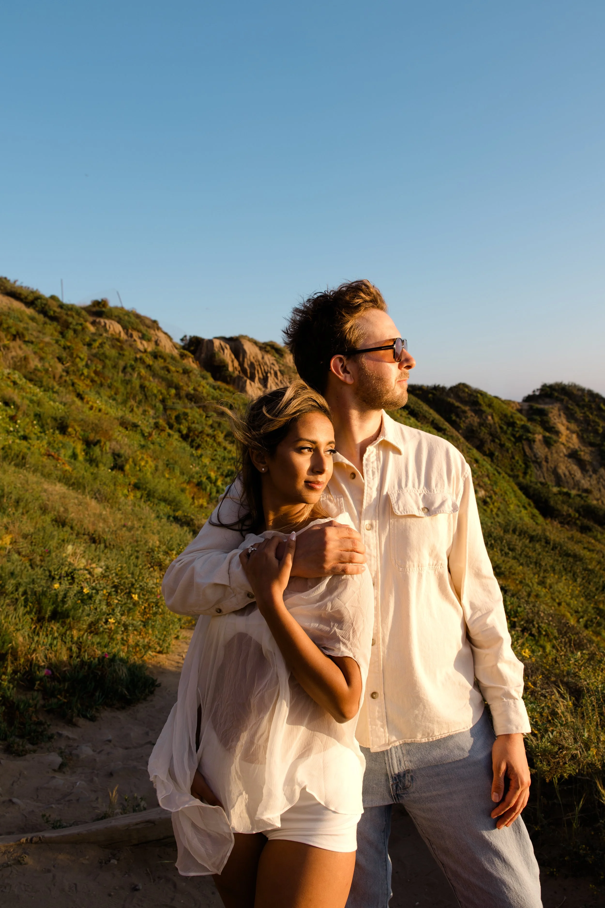 A couple standing outdoors on a hilltop with green vegetation and rocky cliffs in the background, enjoying the sunset, with the woman resting her head on the man's chest.