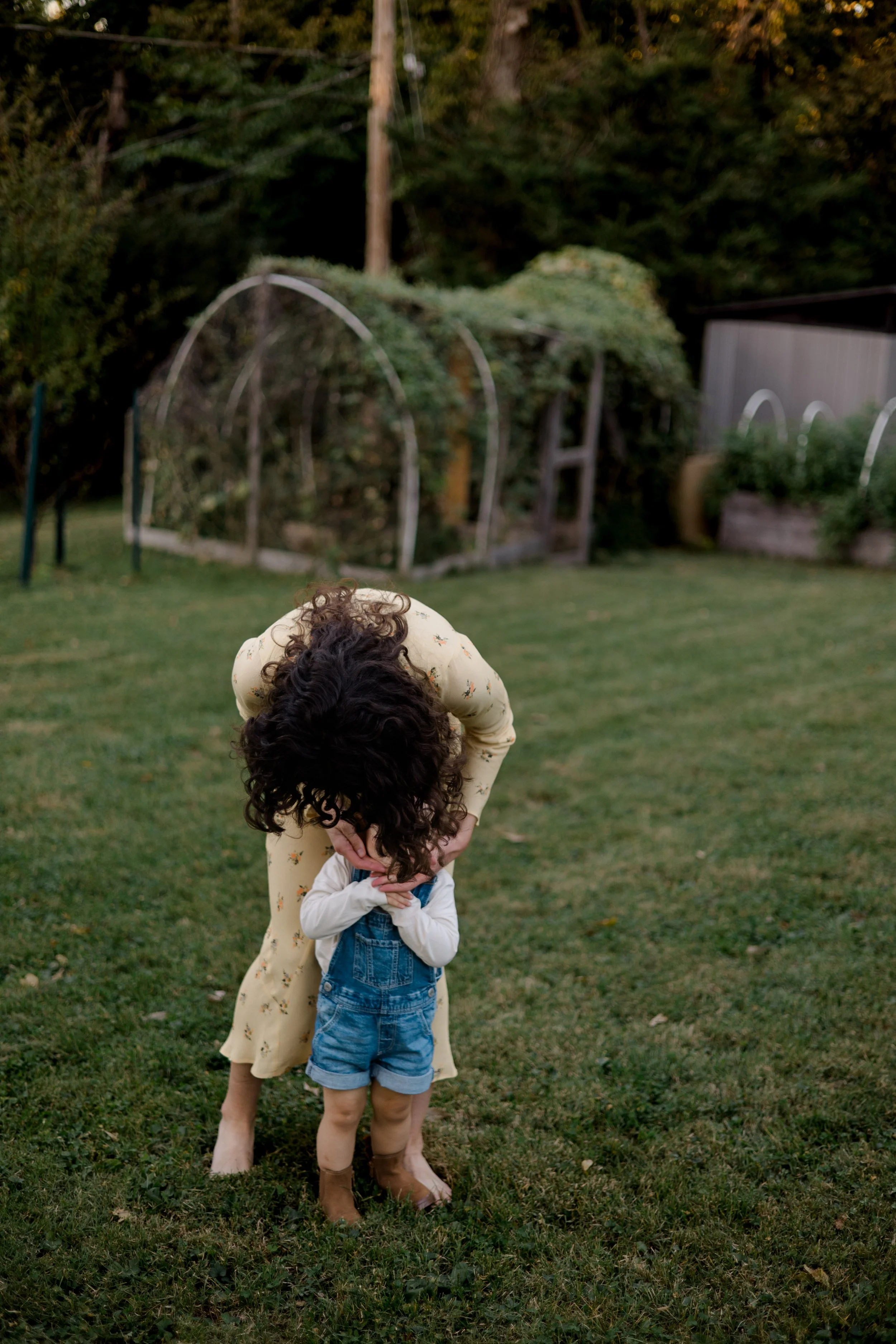 A woman with curly dark hair wearing a yellow dress with small floral patterns is bending over, hugging a small child in a white long-sleeve shirt and denim overalls, in a grassy backyard with trees, bushes, and gardening hoops in the background duri