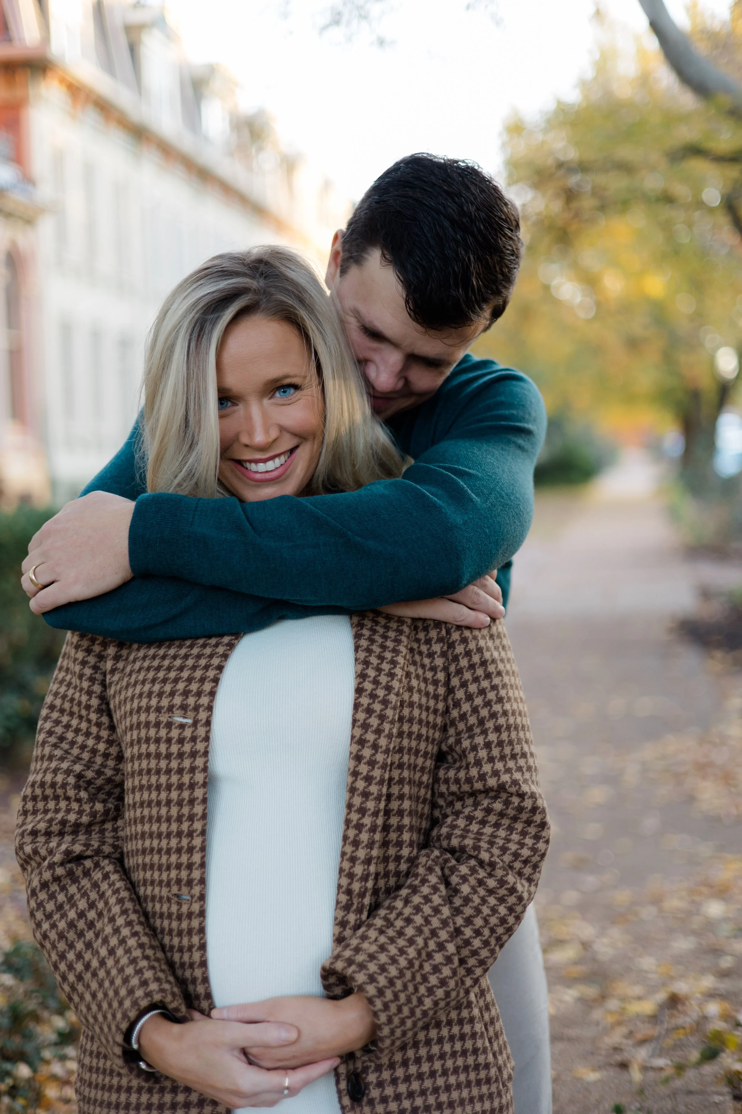 A smiling blonde woman in a brown houndstooth coat standing outdoors with a man hugging her from behind during autumn, with trees and fallen leaves in the background.