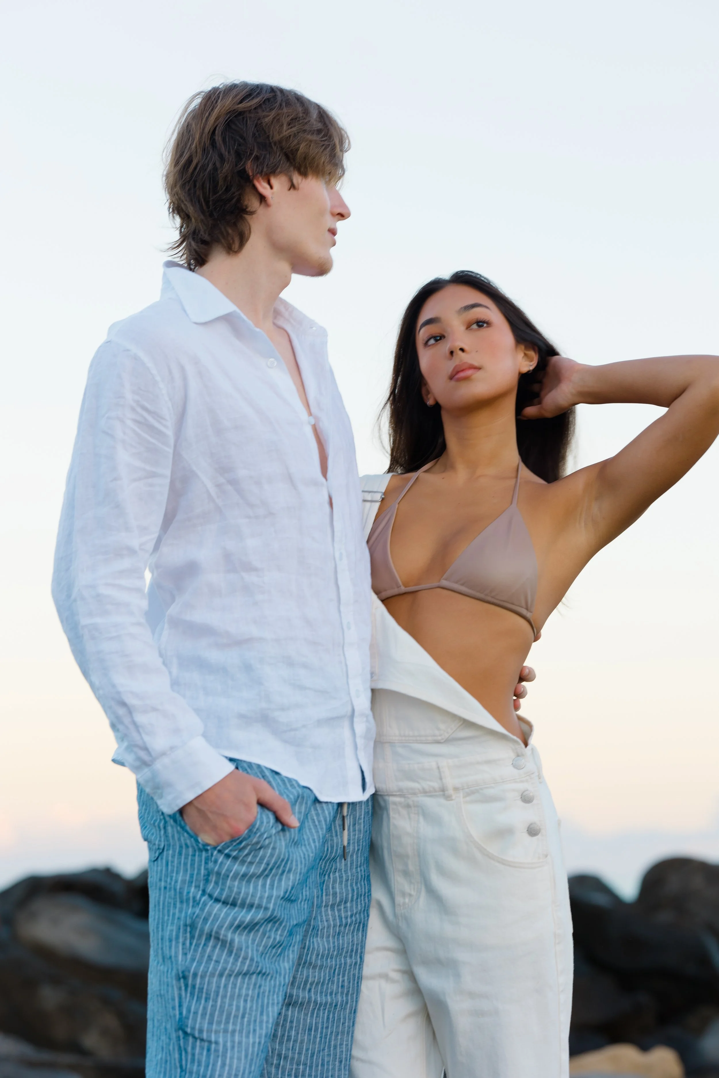 A young man and woman standing outdoors on rocks, looking moody, with a clear sky in the background.