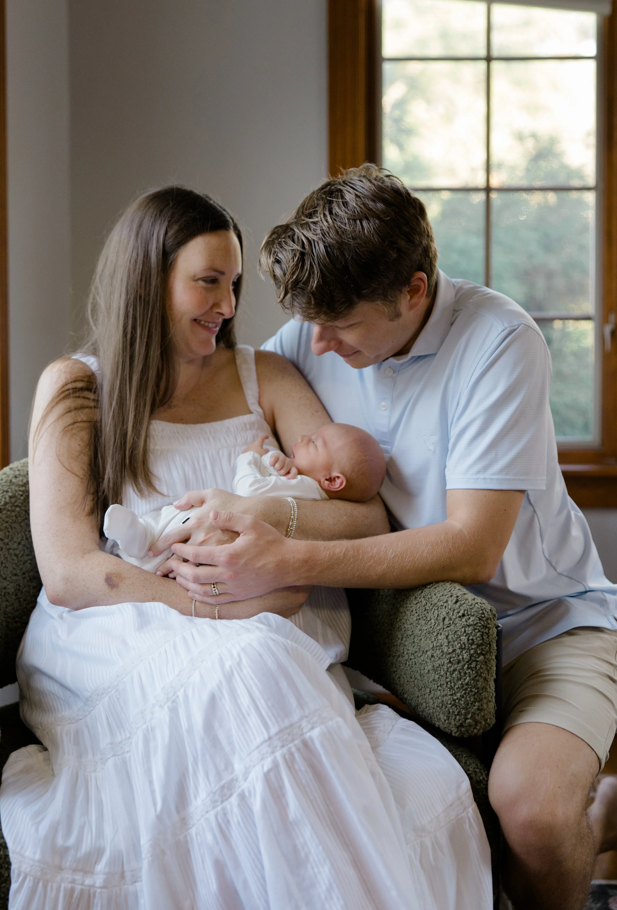 A family with a mother, father, and newborn baby in a cozy home setting, with big windows in the background.