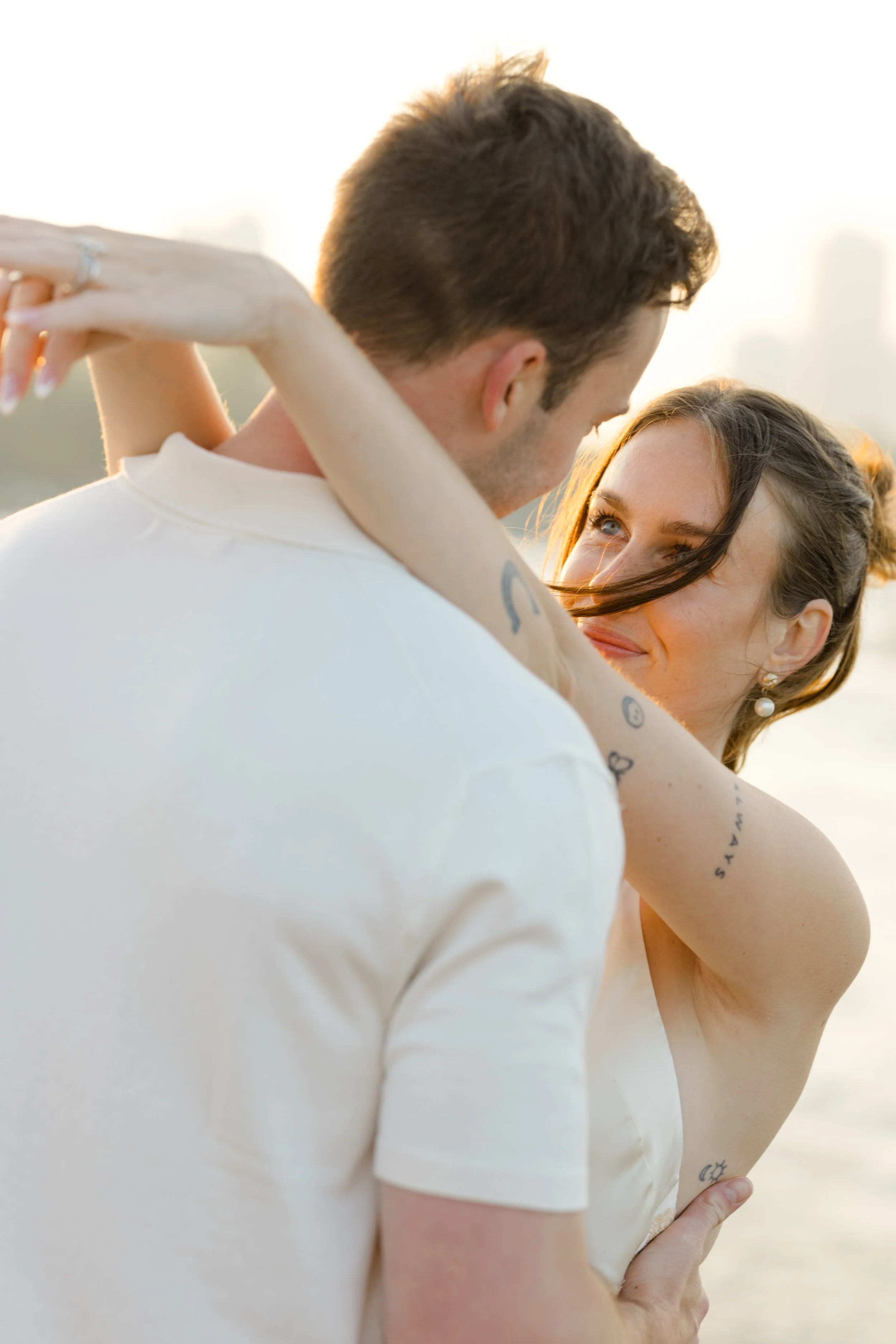 A man and woman dance closely outdoors during sunset, with city buildings faintly visible in the background.