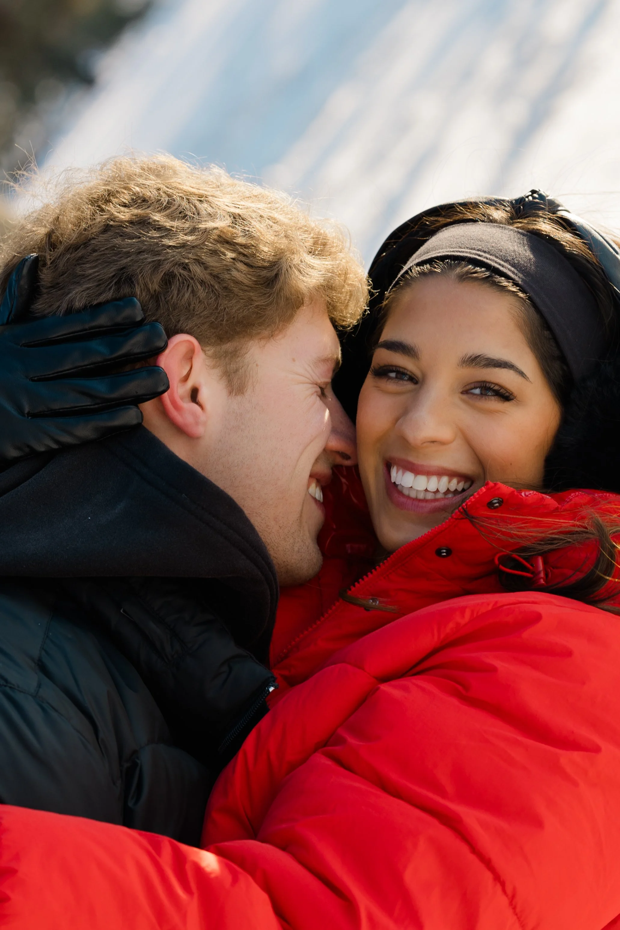 Two smiling people hugging, one with blonde hair and black gloves, the other with dark hair and a black headband, wearing red and black winter jackets, outdoors in the snow.