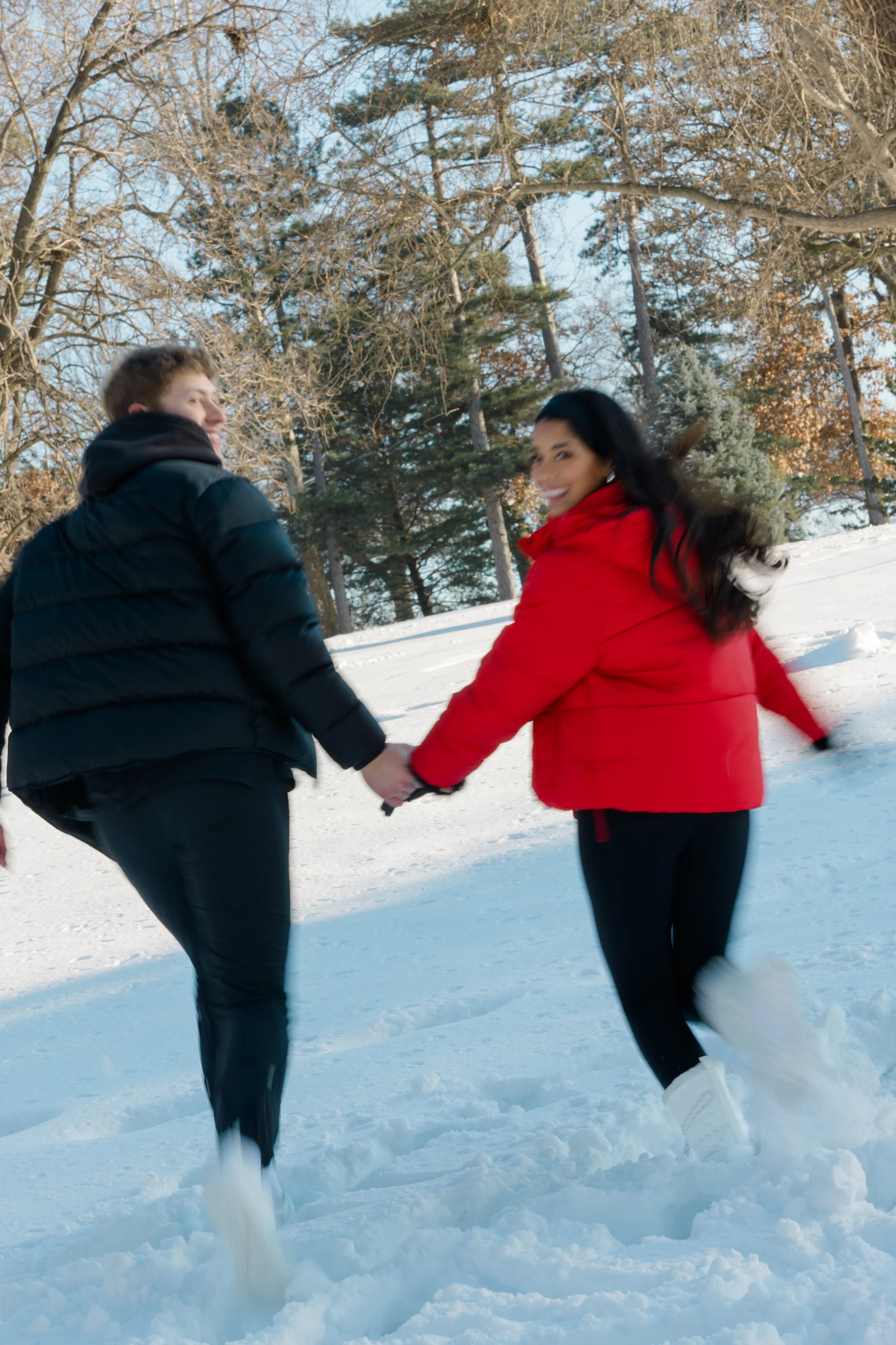 Two people holding hands and spinning in a snowy outdoor setting with snow-covered ground and trees in the background.