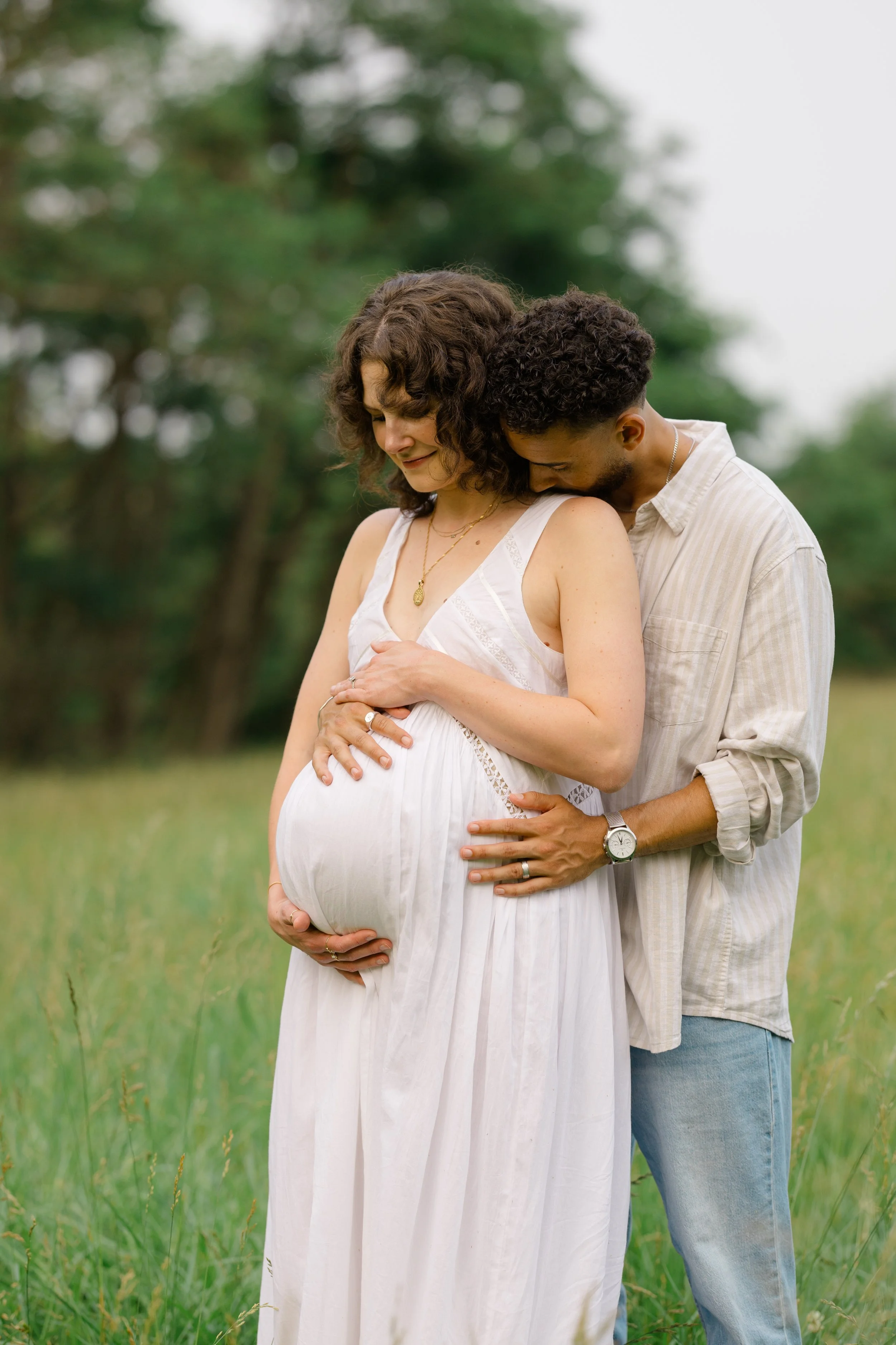 A pregnant woman in a white dress is embracing her partner from behind in a green outdoor setting, both look happy and affectionate.