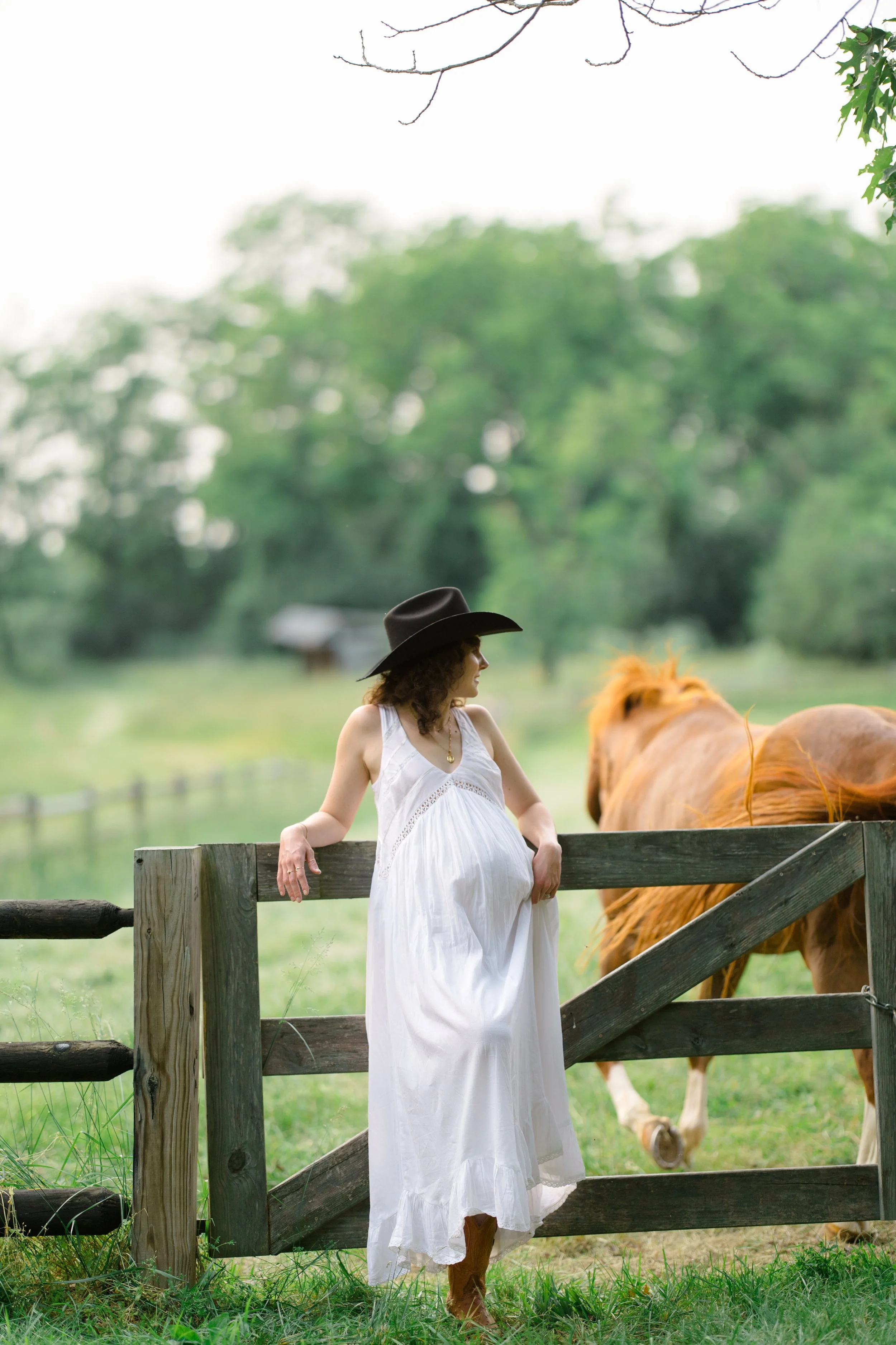 Woman in a white dress and black cowboy hat standing by a wooden fence with a horse grazing nearby on green grass in a rural setting with trees in the background
