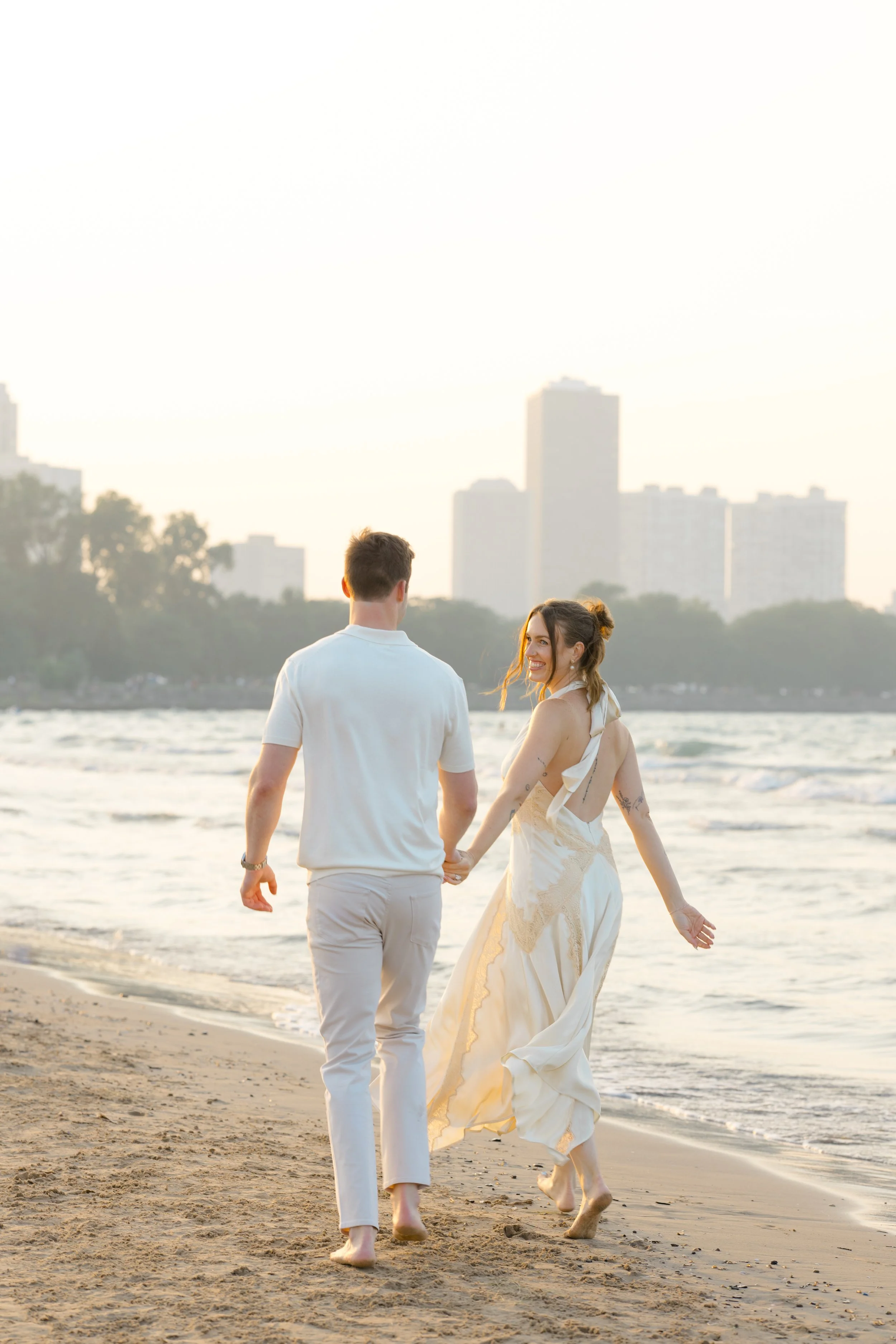A man and woman holding hands and walking barefoot along the beach, wearing light-colored clothing, with city buildings in the background during sunset.
