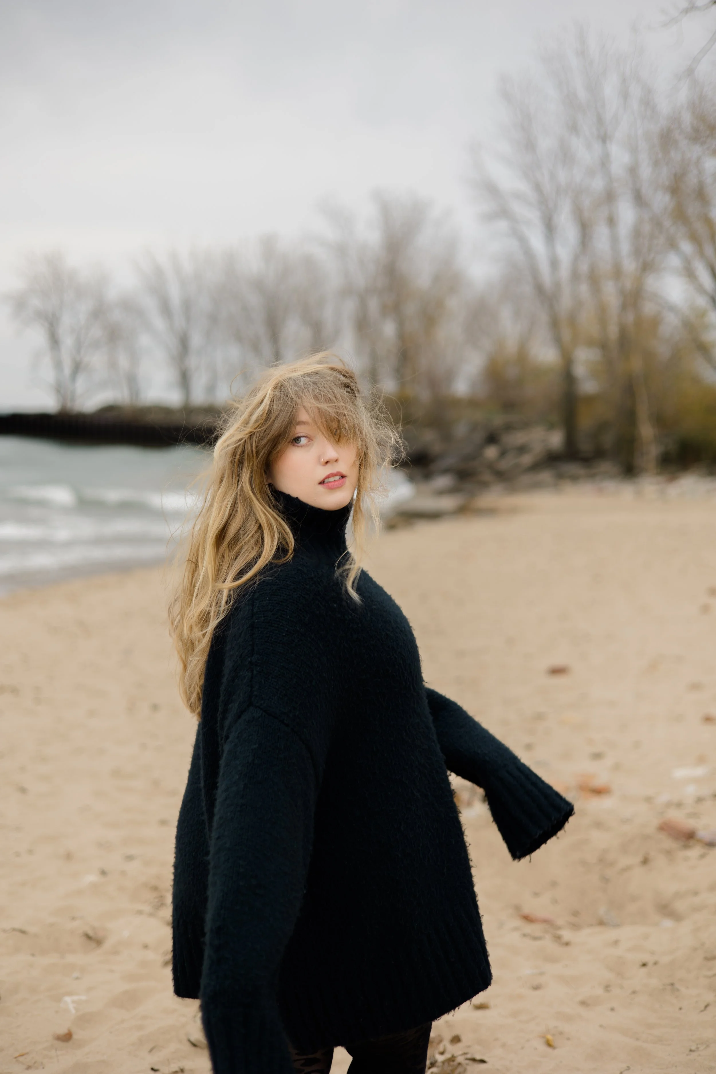 A young woman with wavy blonde hair and a nose piercing sits on a sandy beach, wearing a black turtleneck sweater, with a background of leafless trees and overcast sky.