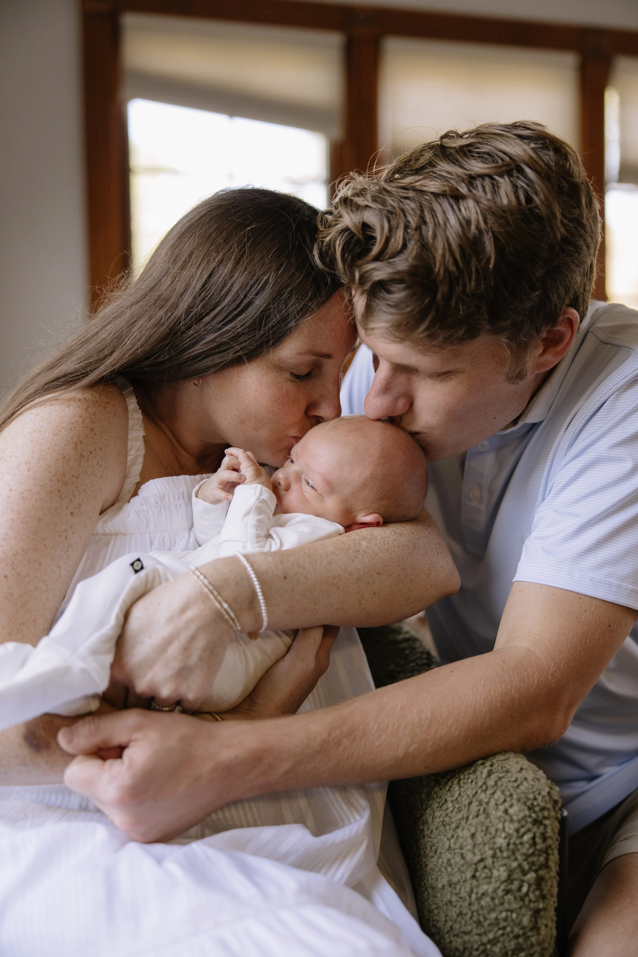 A woman and a man with light skin are kissing their newborn baby, who they are holding in their arms. The woman has long, dark hair and freckles, and the man has short, curly hair. They are indoors near windows with blinds.