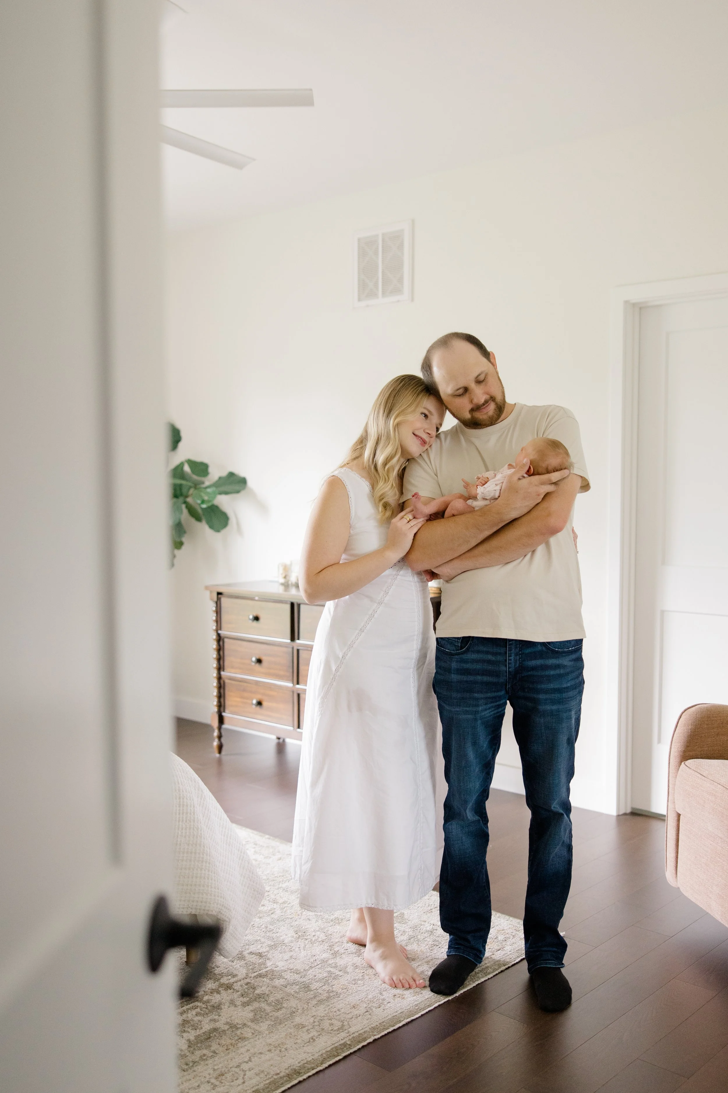 A family with a mother, father, and newborn baby standing together in a bright, modern home, smiling and holding the baby.