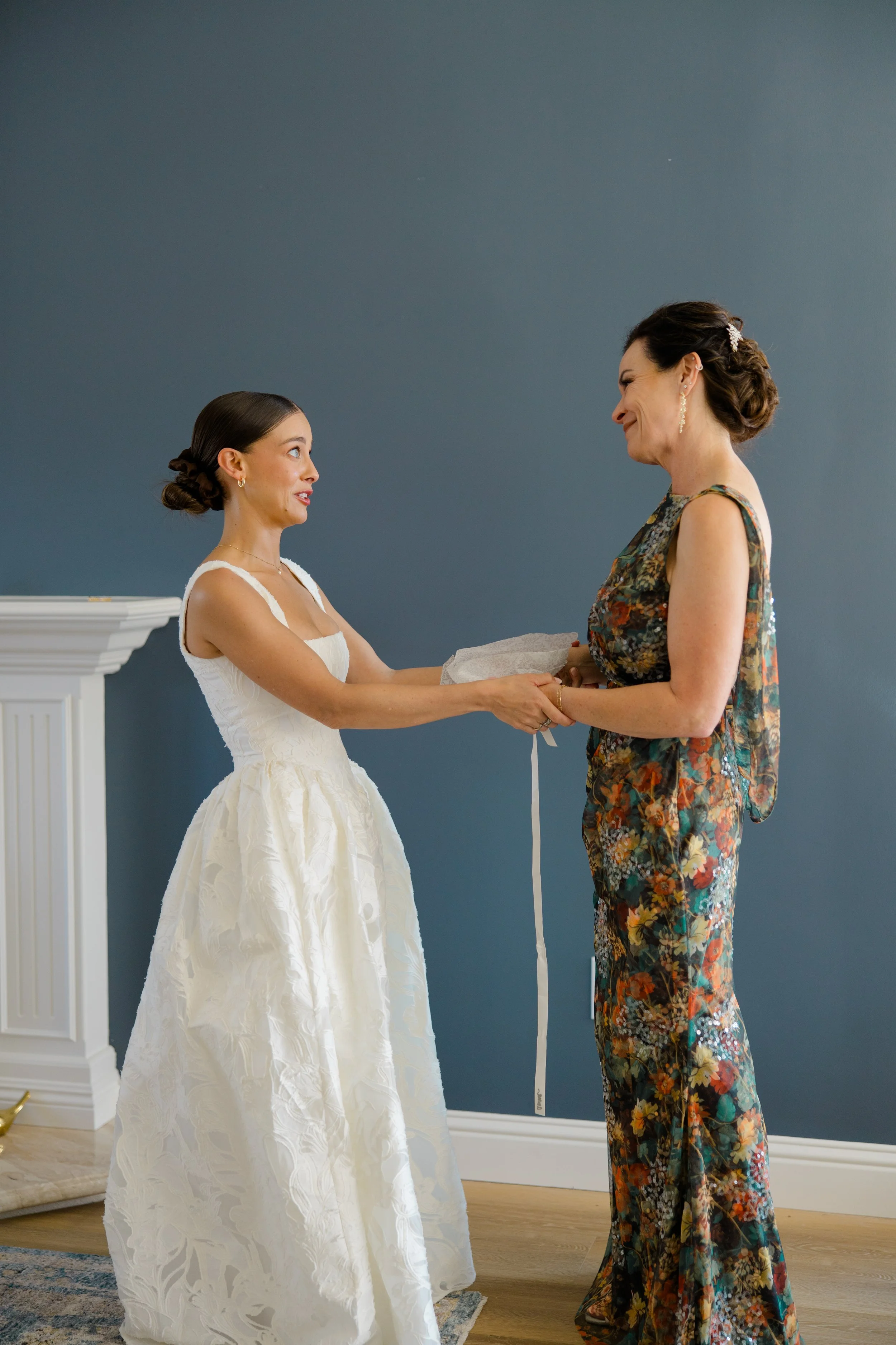 A bride and an older woman holding hands, facing each other indoors against a blue wall. The bride wears a white wedding dress, and the older woman wears a multicolored floral dress with earrings and a hair accessory. They are smiling and looking at 