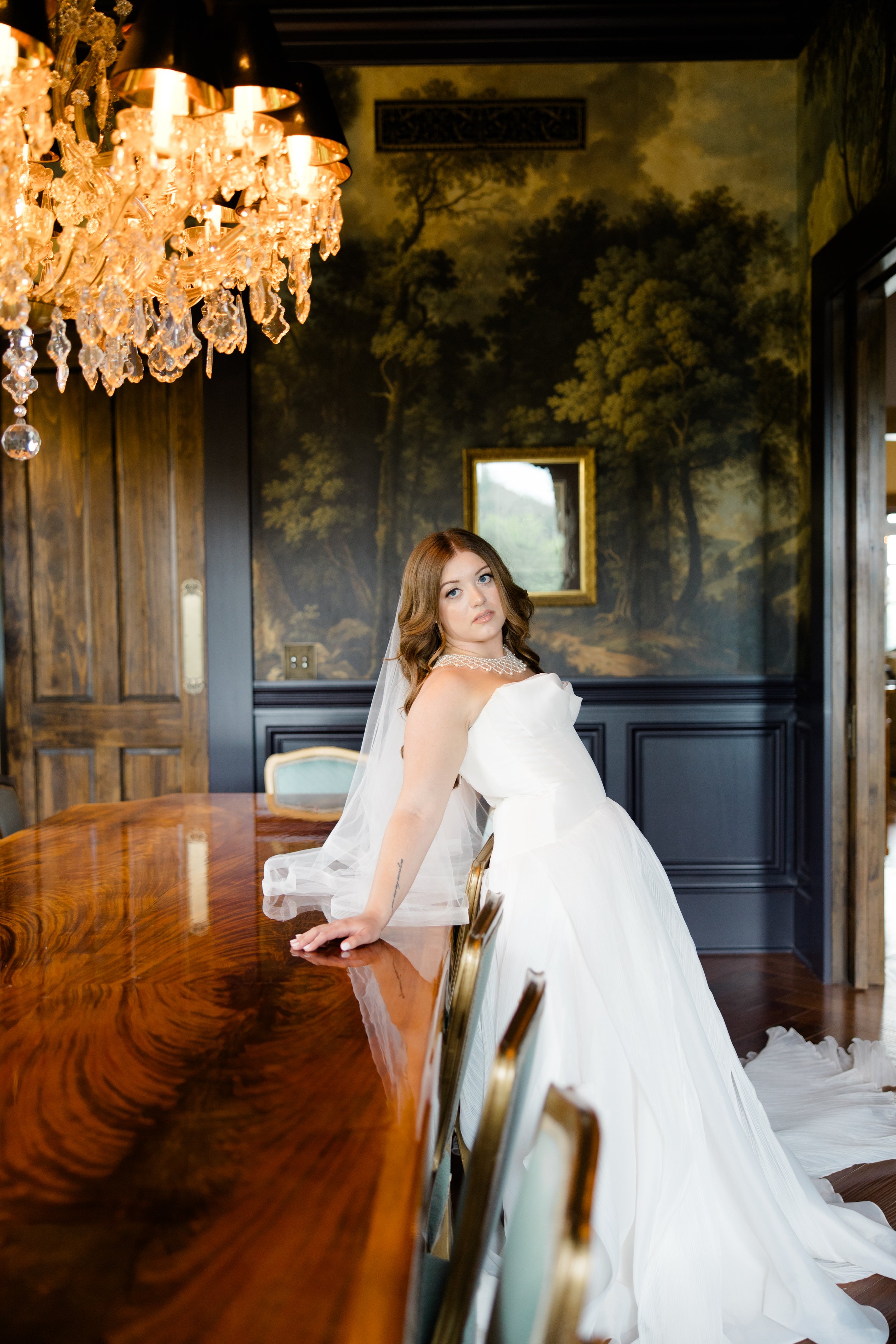A bride with shoulder-length brown hair in a white wedding gown with off-shoulder design and pearl necklace, leaning on a polished wooden table in a decorated elegant room with a chandelier, painting, and dark wood paneling.