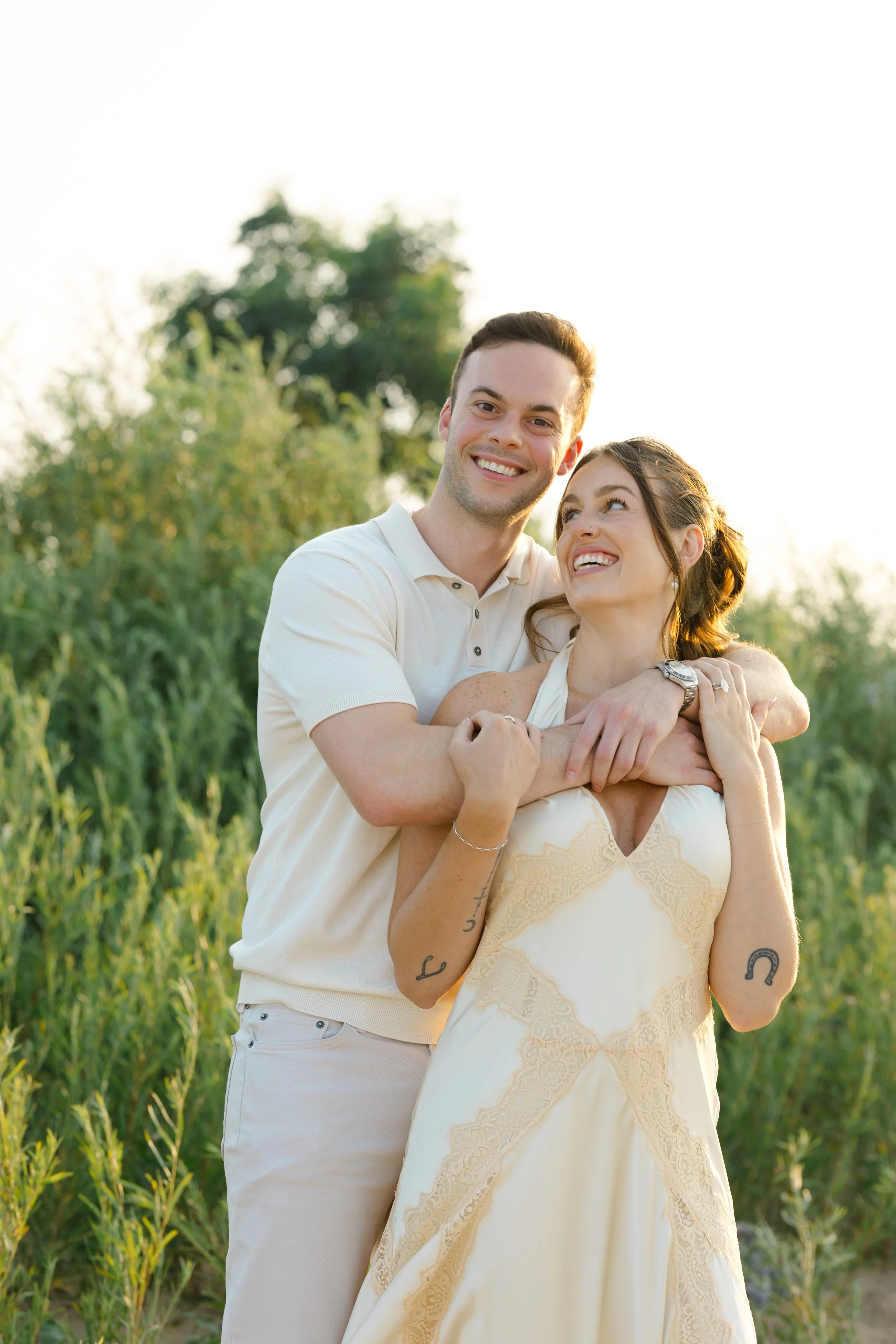 A happy couple stands outdoors in a green field, with tall grass and trees in the background, embracing each other and smiling at the camera during golden hour.